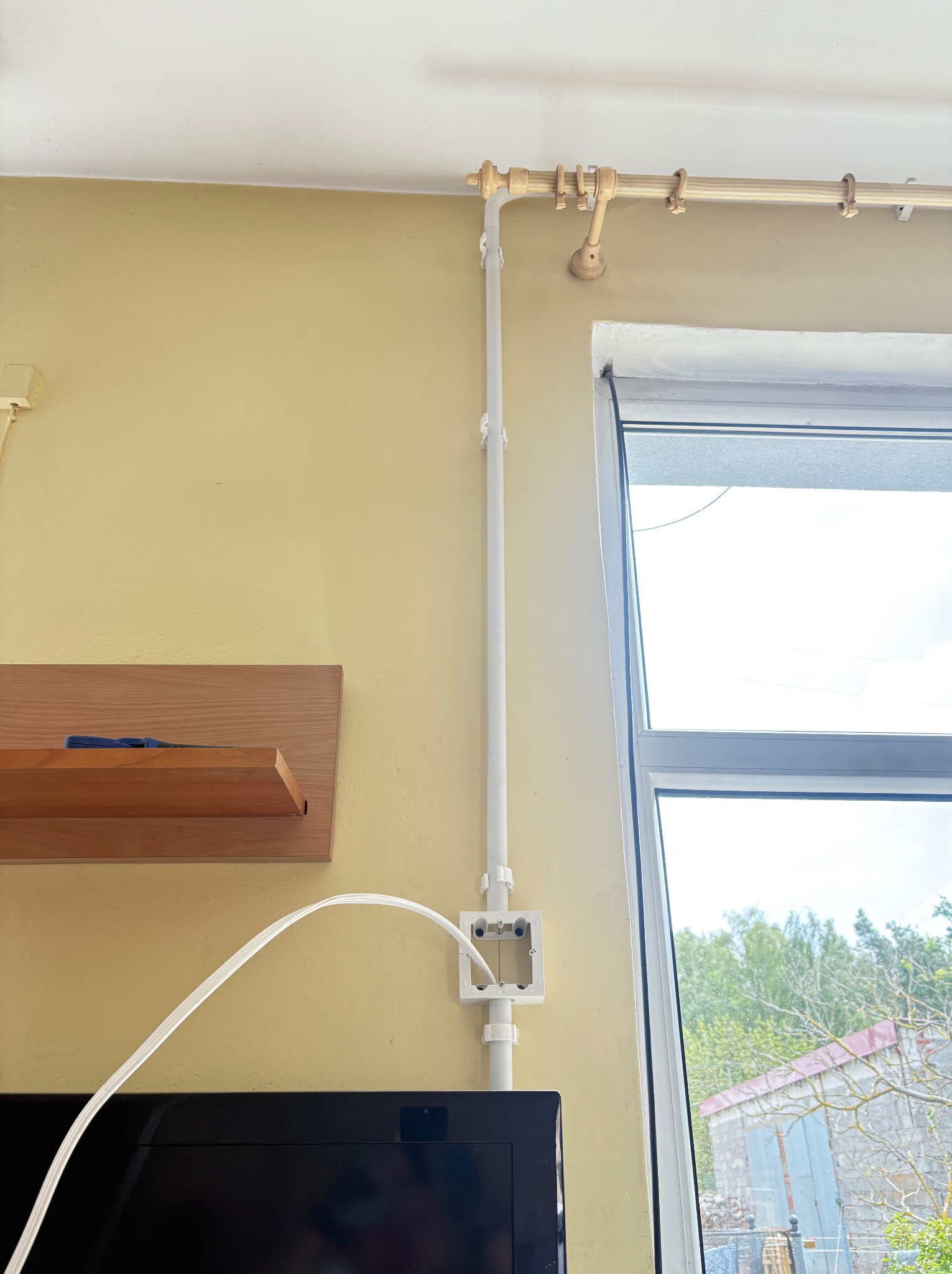 Biała puszka elektryczna natynkowa z kablem w żółtym pokoju, widok z dołu. Rura osłonowa pionowo do sufitu, obok okna. Fragment półki i telewizora.