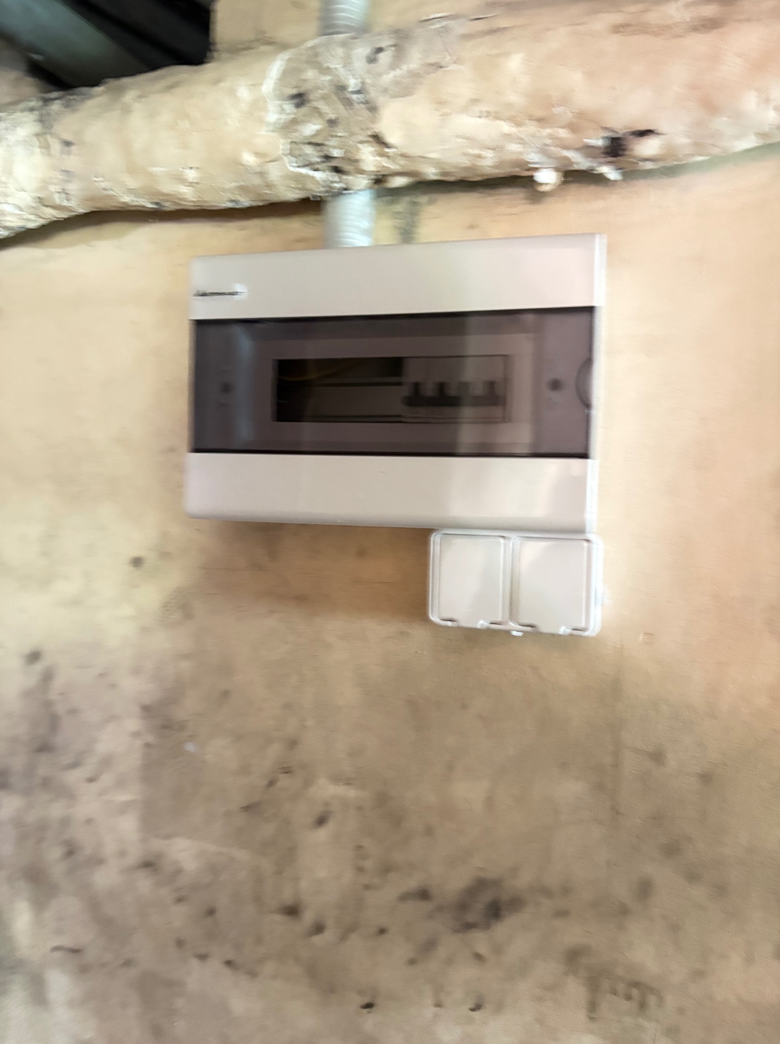 Szara skrzynka elektryczna z bezpiecznikami, zamontowana na ścianie z rurą w tle. Widoczne zabezpieczenia i obudowa z tworzywa.