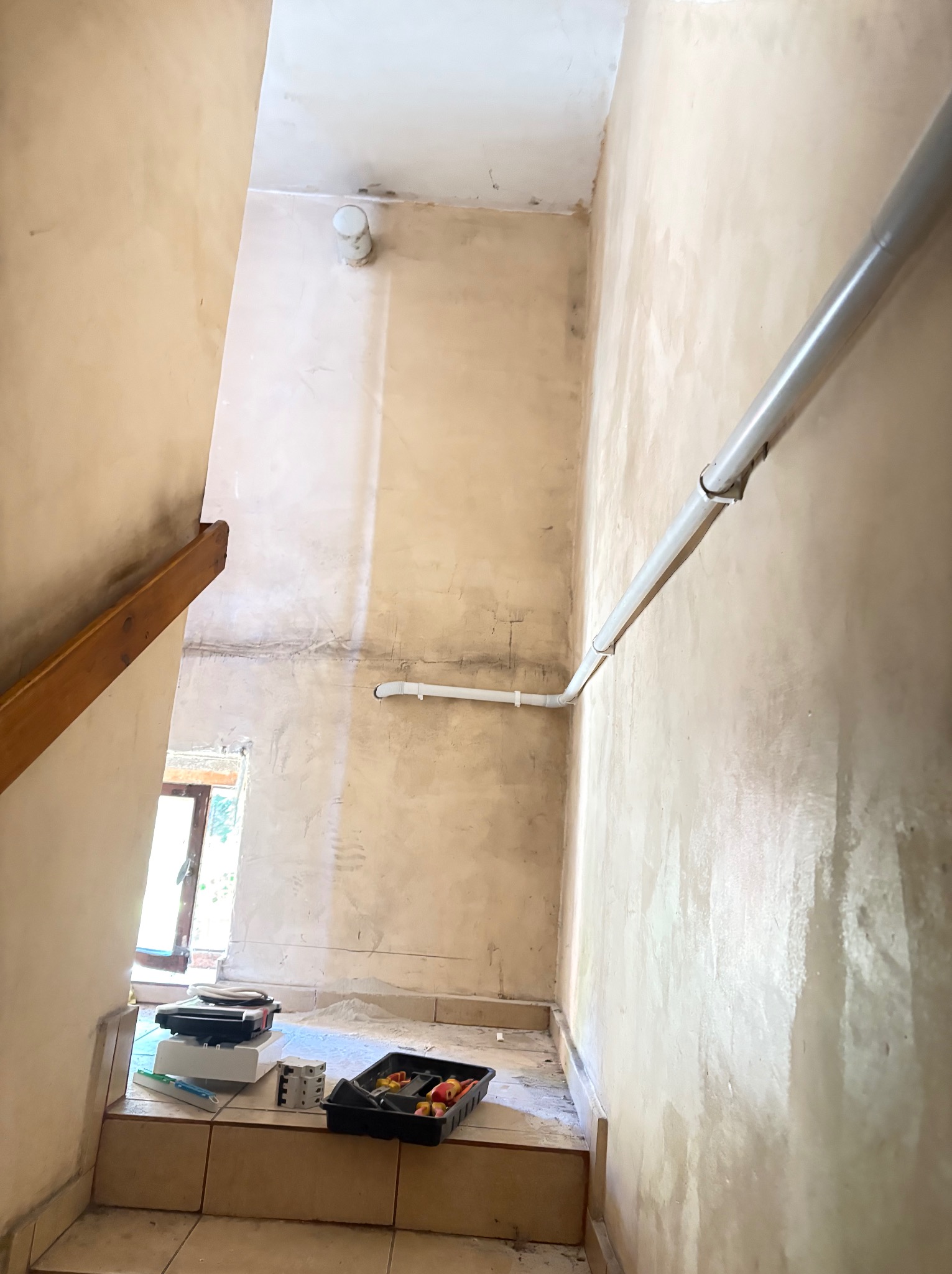 Klatka schodowa w trakcie remontu. Widoczne narzędzia malarskie, ściany po wstępnym malowaniu, rury instalacyjne. Naturalne światło wpada przez okno.
