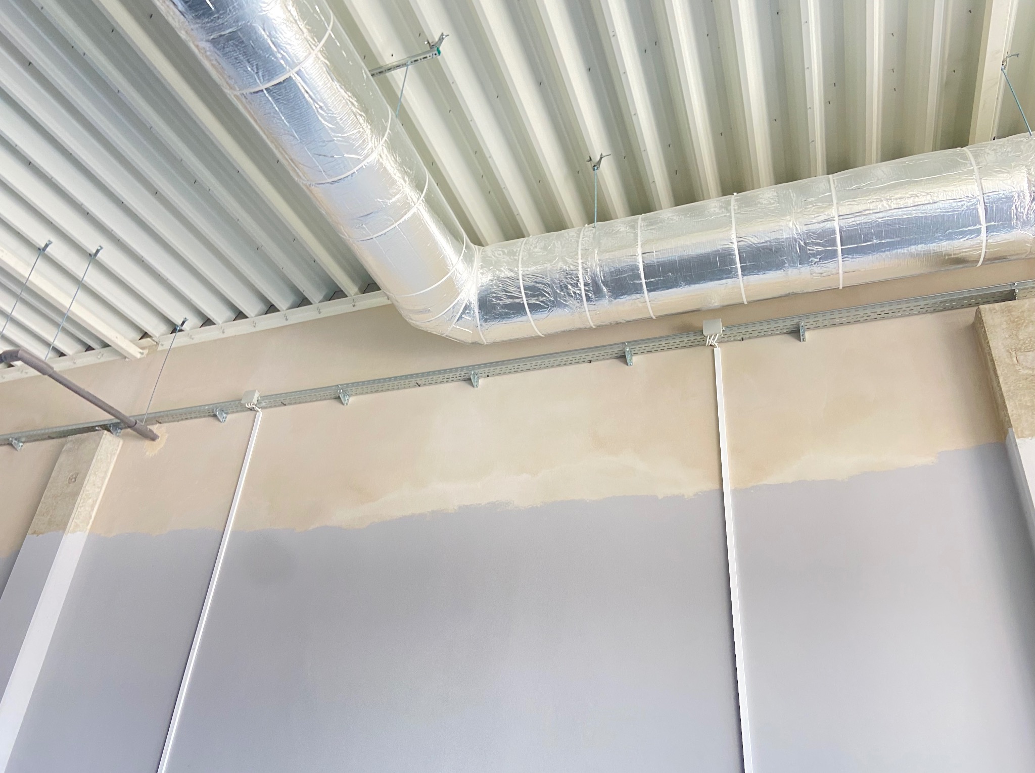 Szara ściana z korytem kablowym i rurą wentylacyjną pod sufitem z blachy trapezowej. Instalacja widoczna w trakcie prac wykończeniowych.