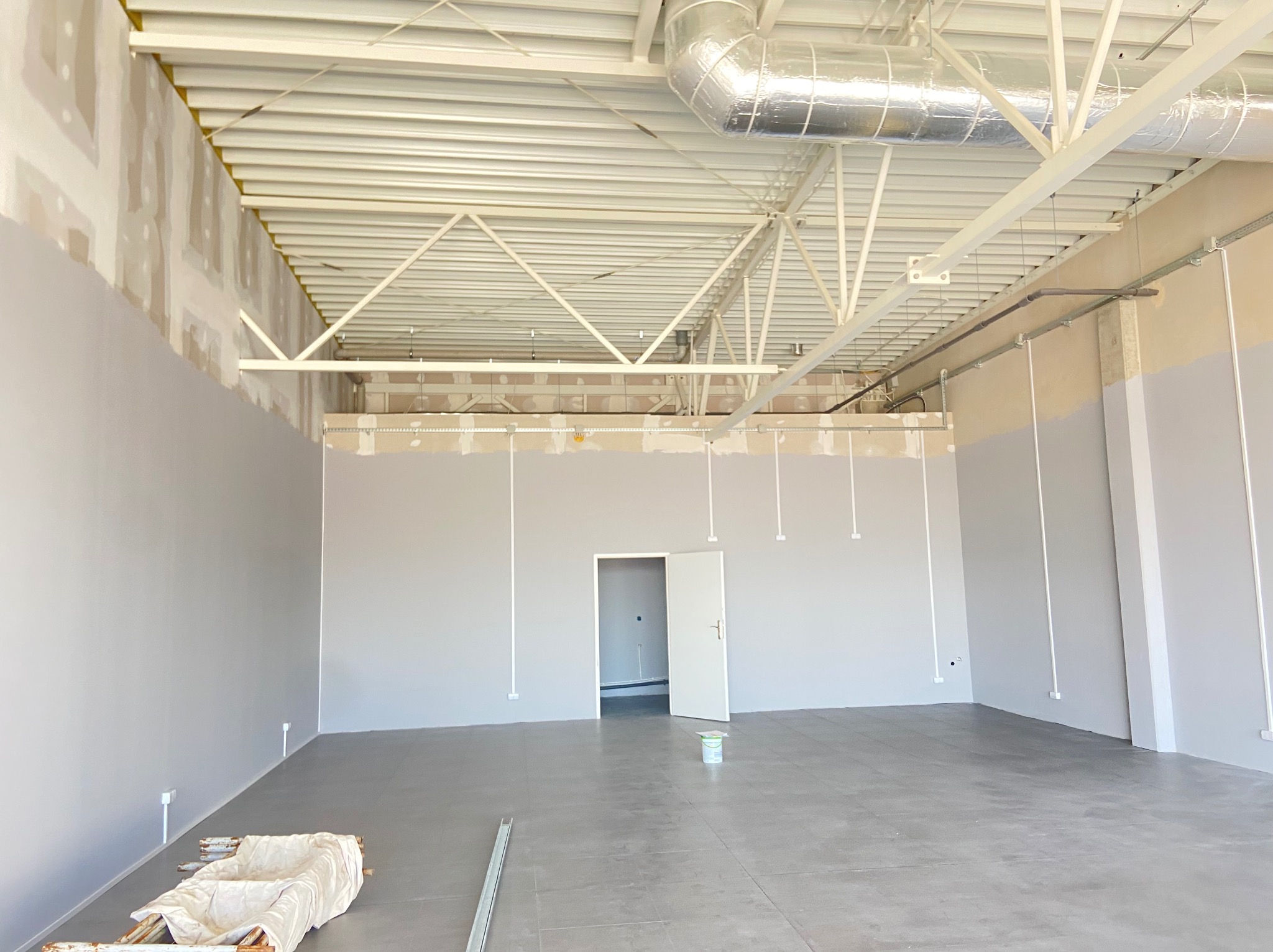 Wnętrze w trakcie prac wykończeniowych: szpachlowane ściany, widoczne kable elektryczne w peszlach, surowa podłoga. Hala z metalową konstrukcją dachu.