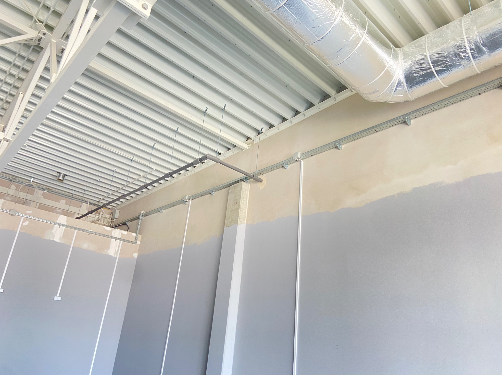 Kable w korytkach i rury pod sufitem w nowym budynku. Szare ściany w połowie pomalowane. Widok na sufit z blachy trapezowej i konstrukcję stalową.