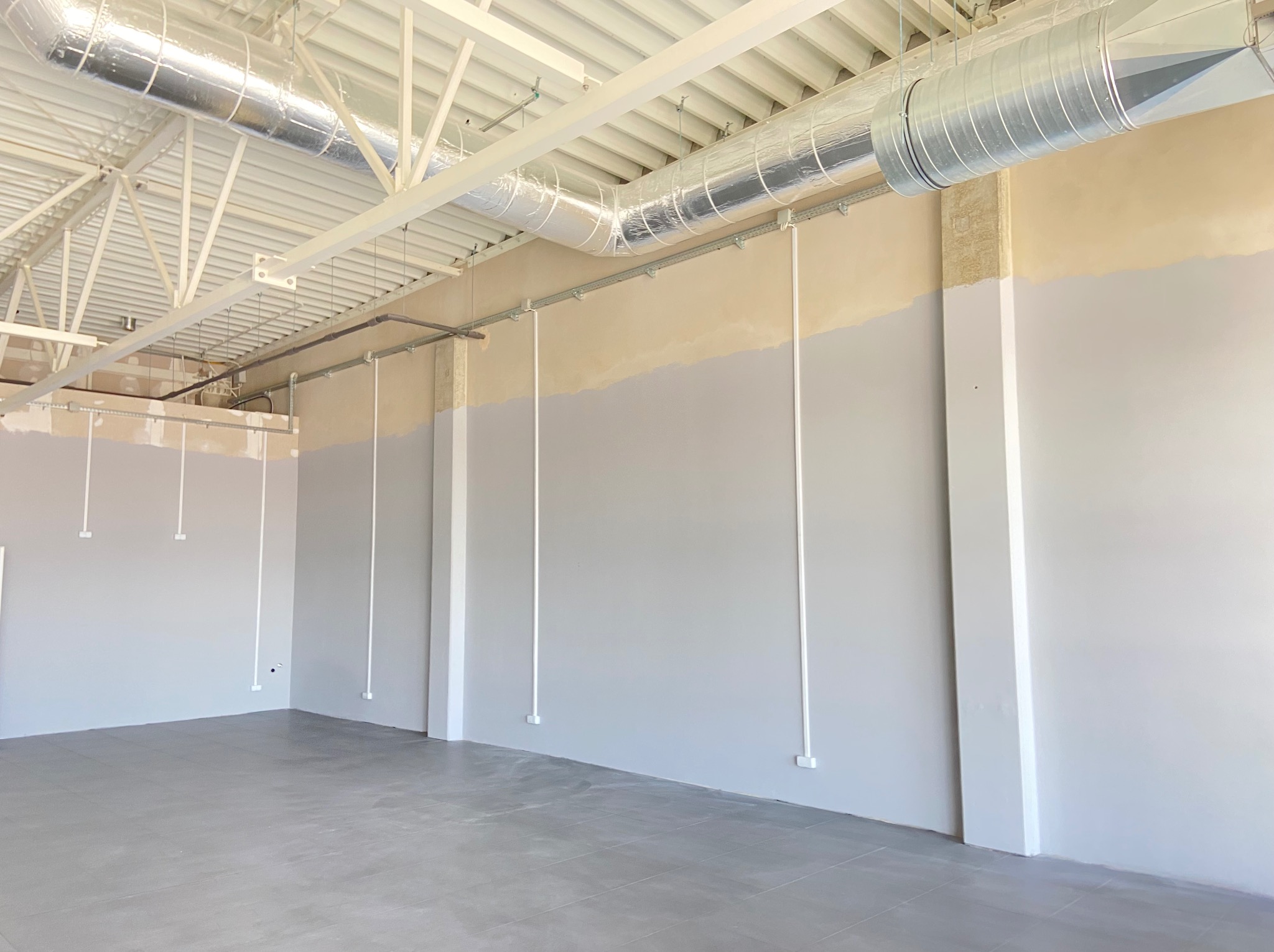 Surowe wnętrze: szara ściana, podłoga z płytek, rury wentylacyjne pod sufitem. Widoczne kable i puszki elektryczne na ścianie w trakcie instalacji.