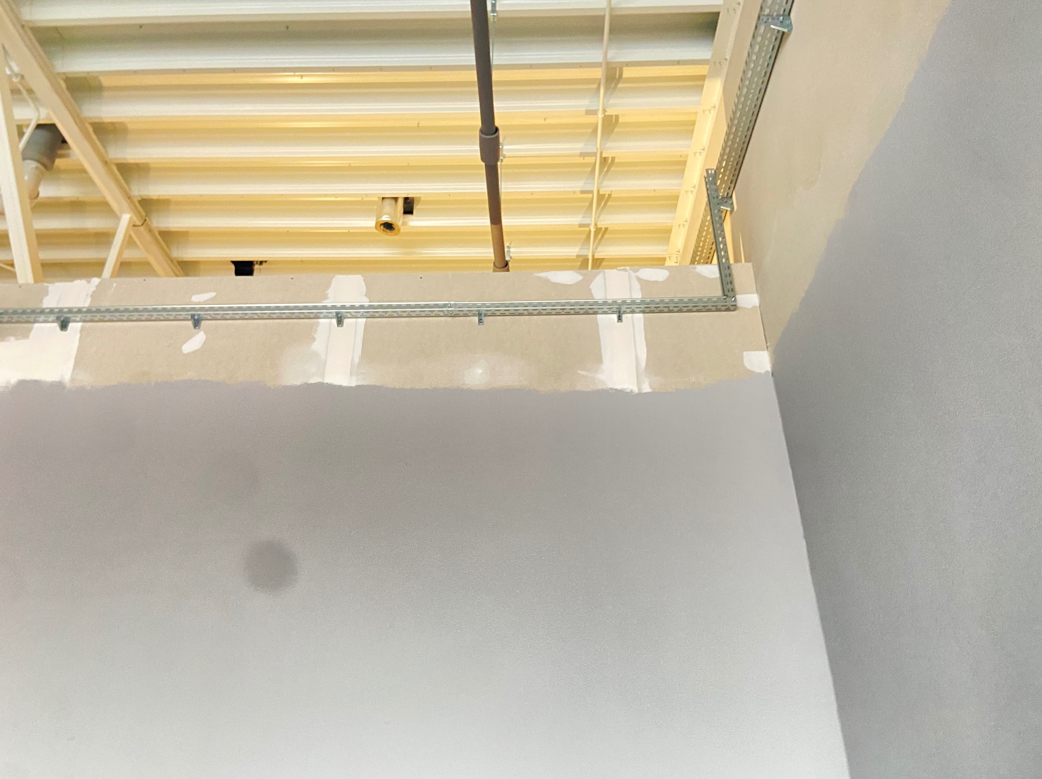 Szara ściana z metalowymi elementami mocującymi pod sufitem. Widoczne rury i kable pod stropem. Prace wykończeniowe wewnątrz budynku.