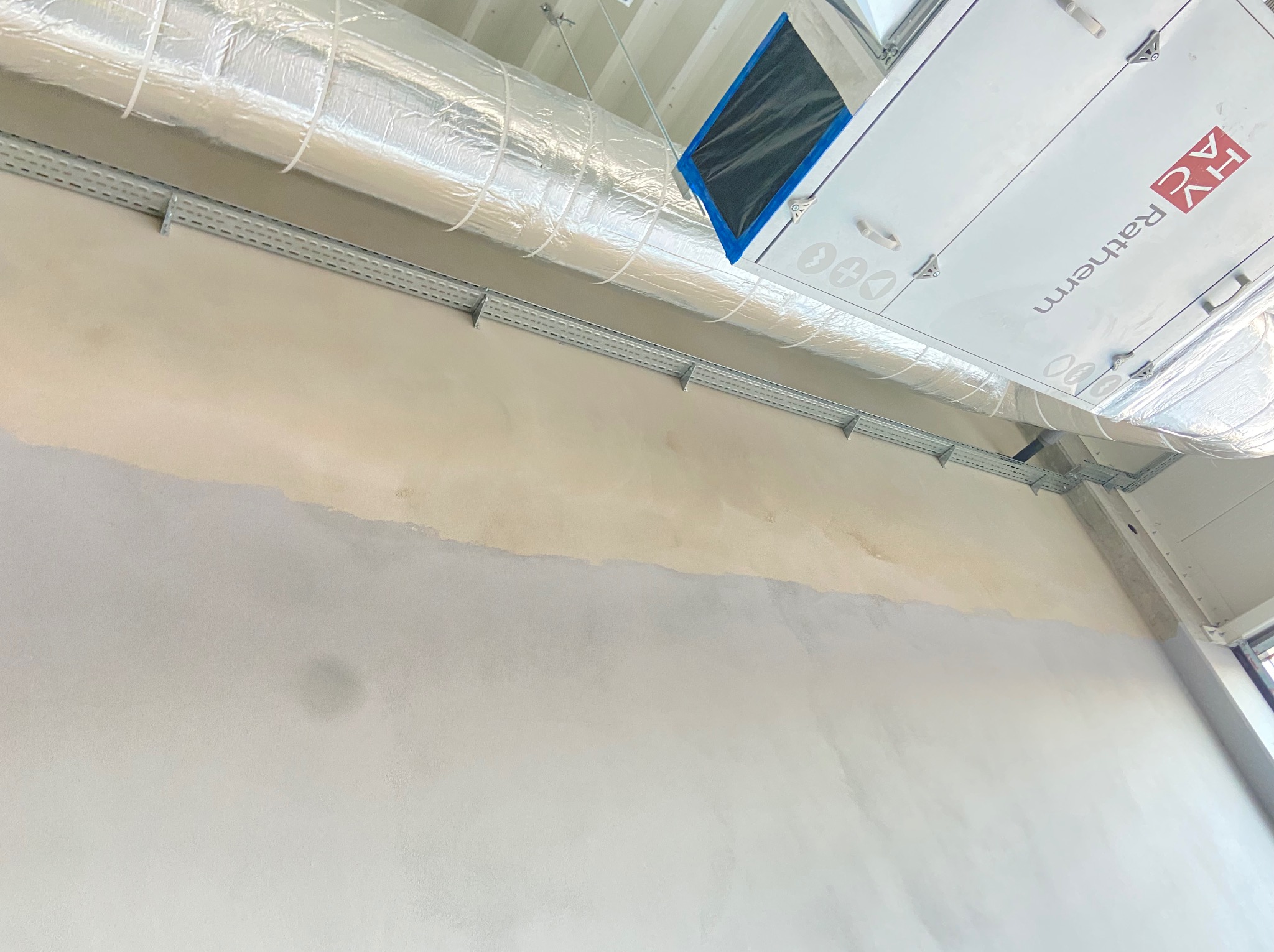 Metalowe korytka kablowe zamontowane na ścianie wewnątrz budynku, widoczne elementy instalacji wentylacyjnej pod sufitem.