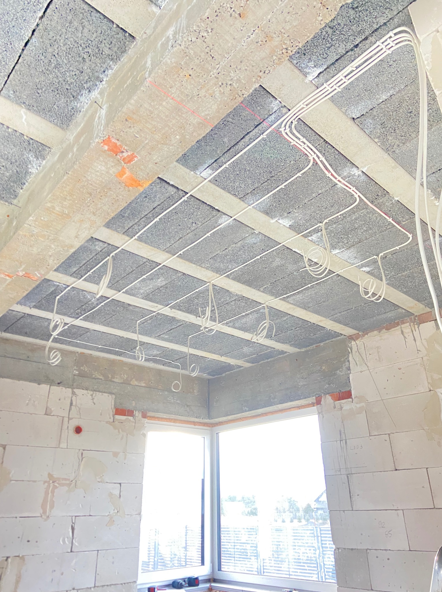 Białe kable elektryczne ułożone pod betonowym sufitem w surowym stanie, widoczne elementy konstrukcyjne i okno w tle. Etap prac elektrycznych w nowym budynku.