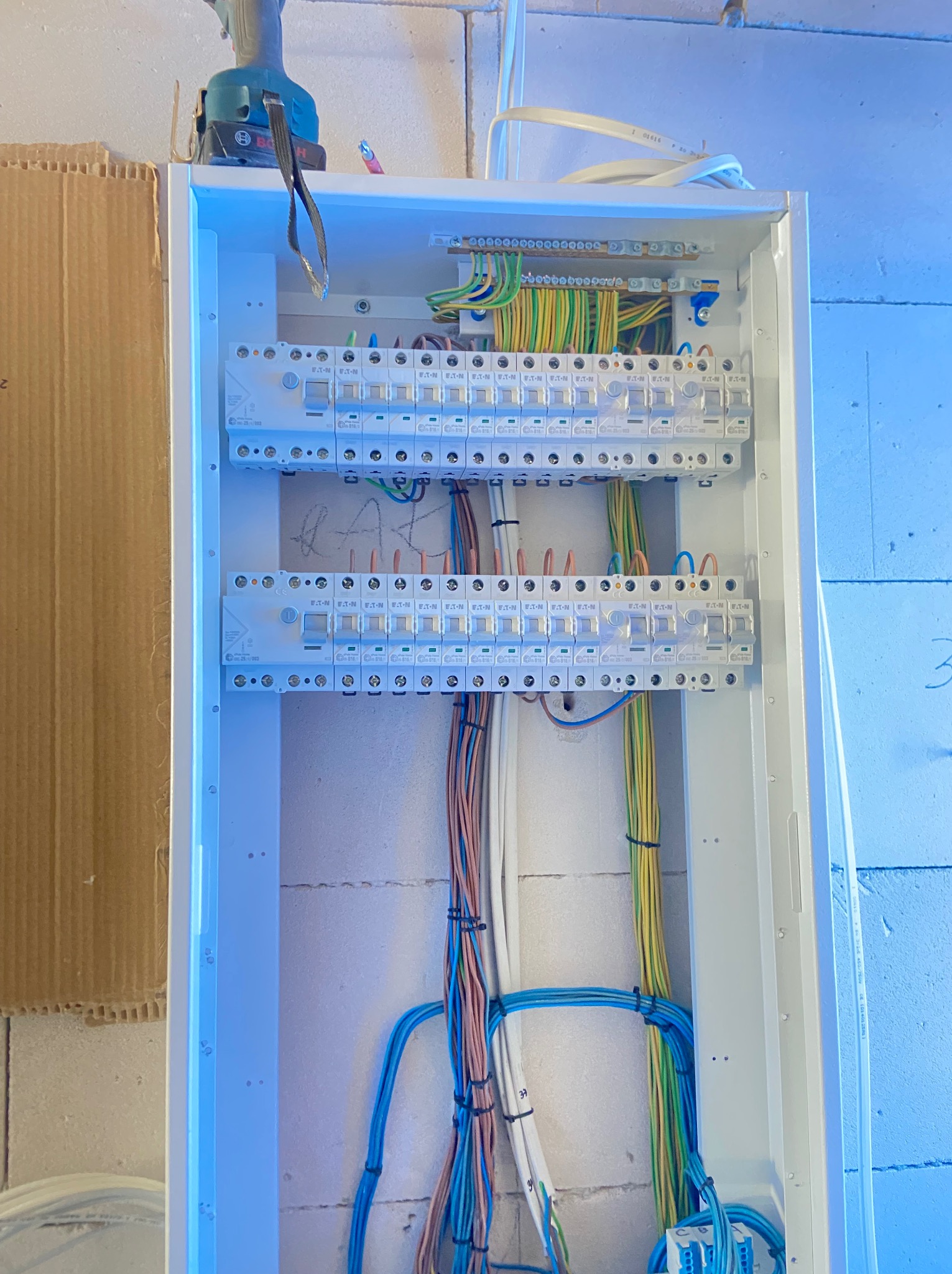 Rozdzielnica elektryczna w trakcie montażu z widocznymi przewodami, bezpiecznikami i narzędziem w tle, na ścianie z bloczków. Instalacje elektryczne wewnątrz budynku.