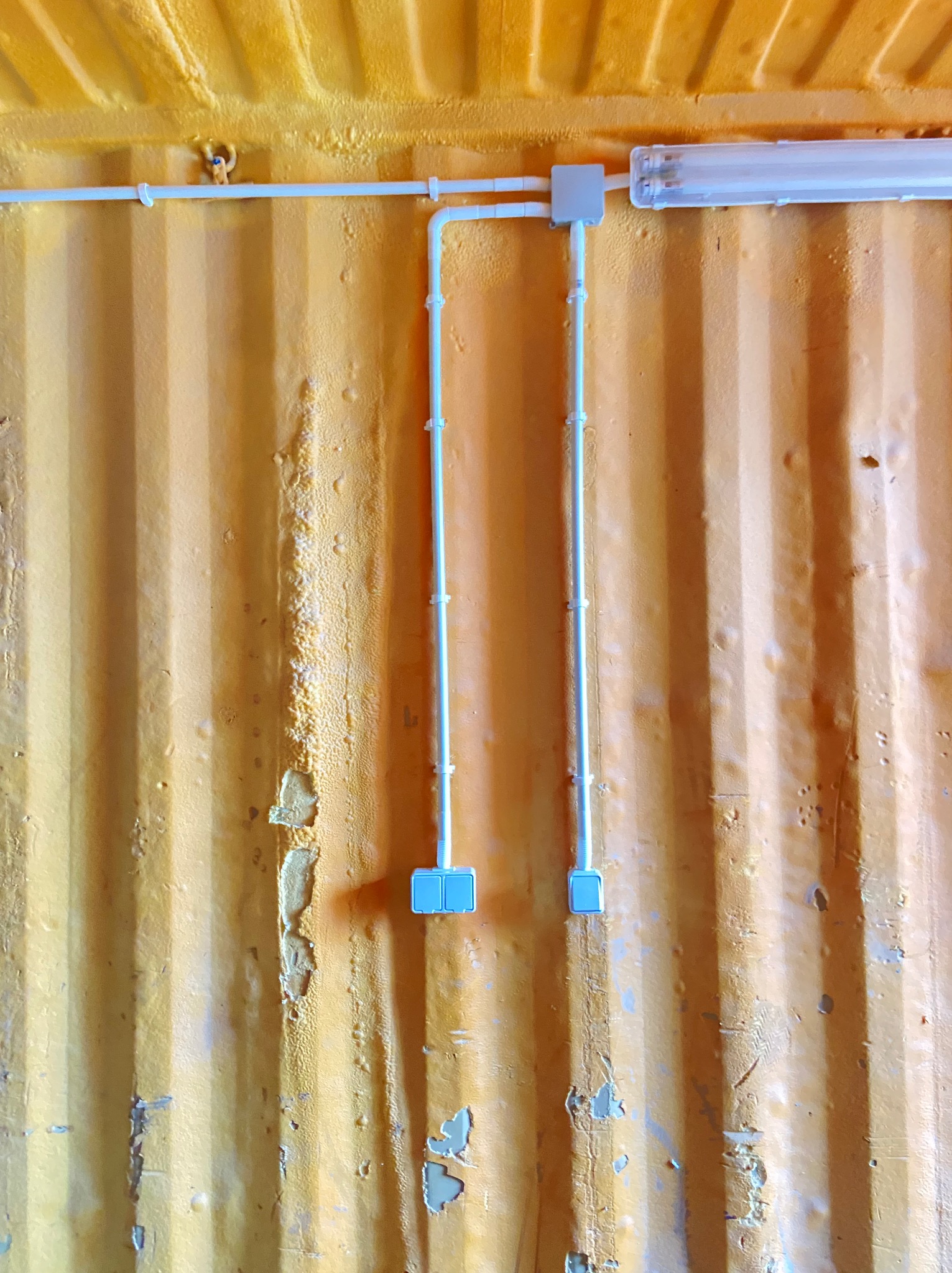 Białe kable elektryczne przymocowane do pomarańczowej, metalowej ściany kontenera z widoczną lampą fluorescencyjną i puszkami instalacyjnymi.