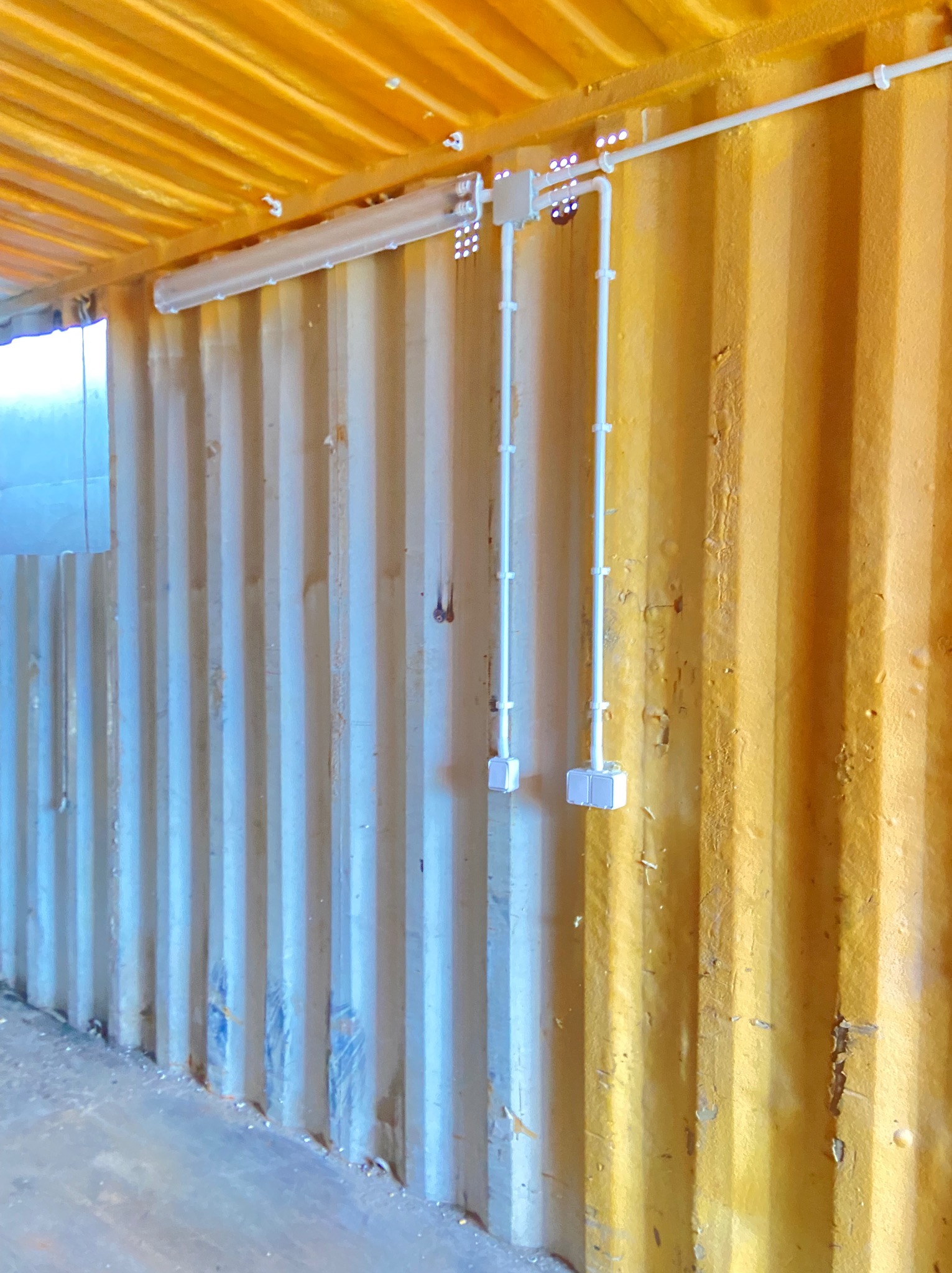 Wnętrze kontenera z żółtymi ścianami, widoczne białe rurki instalacji elektrycznej, gniazdka i świetlówka. Ujęcie z dołu, oświetlenie sztuczne.