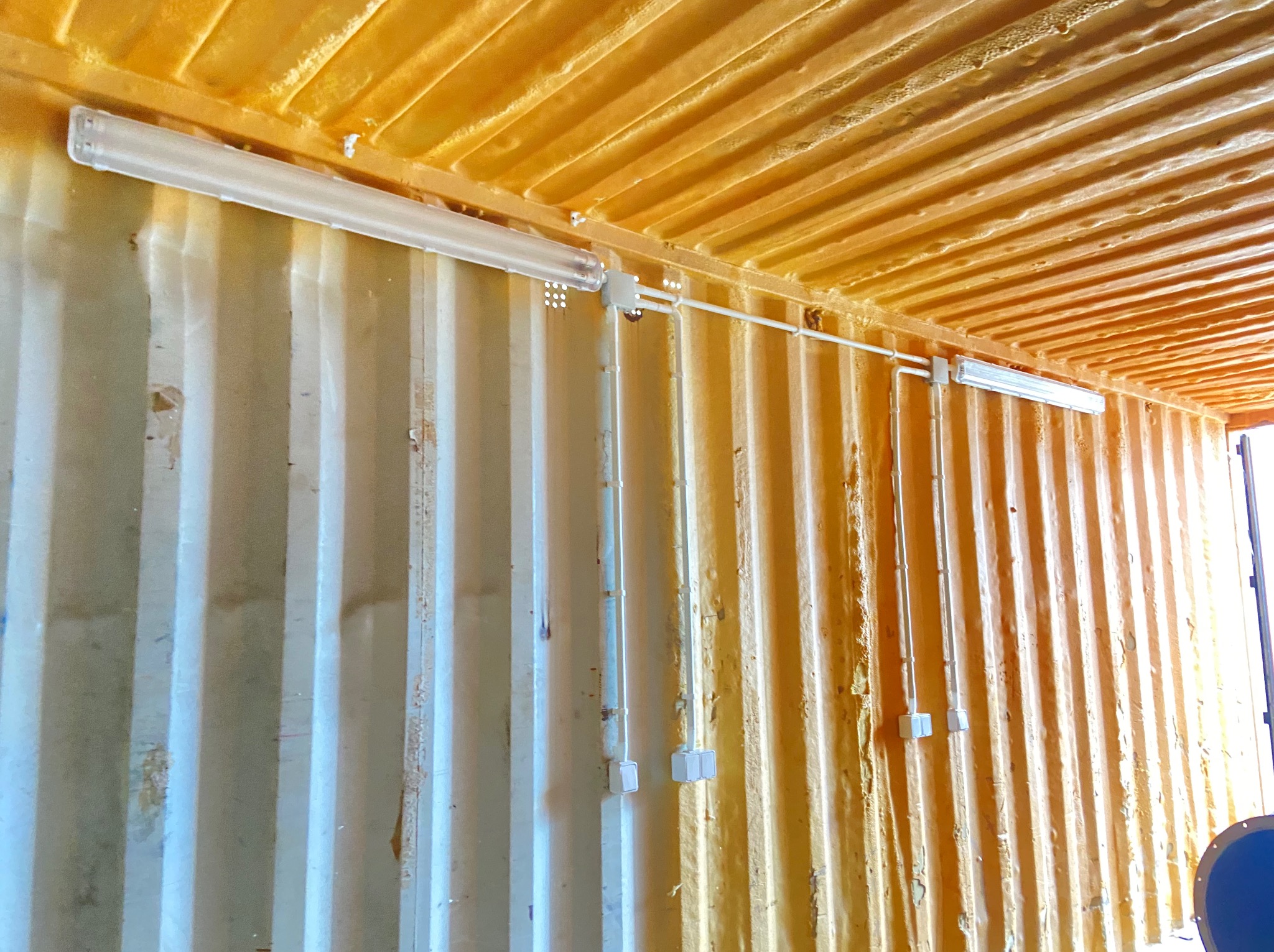 Instalacja oświetlenia w kontenerze morskim: lampy fluorescencyjne zamontowane na ścianie z blachy falistej, przewody w białych peszlach. Sufit ocieplony pianką PUR.