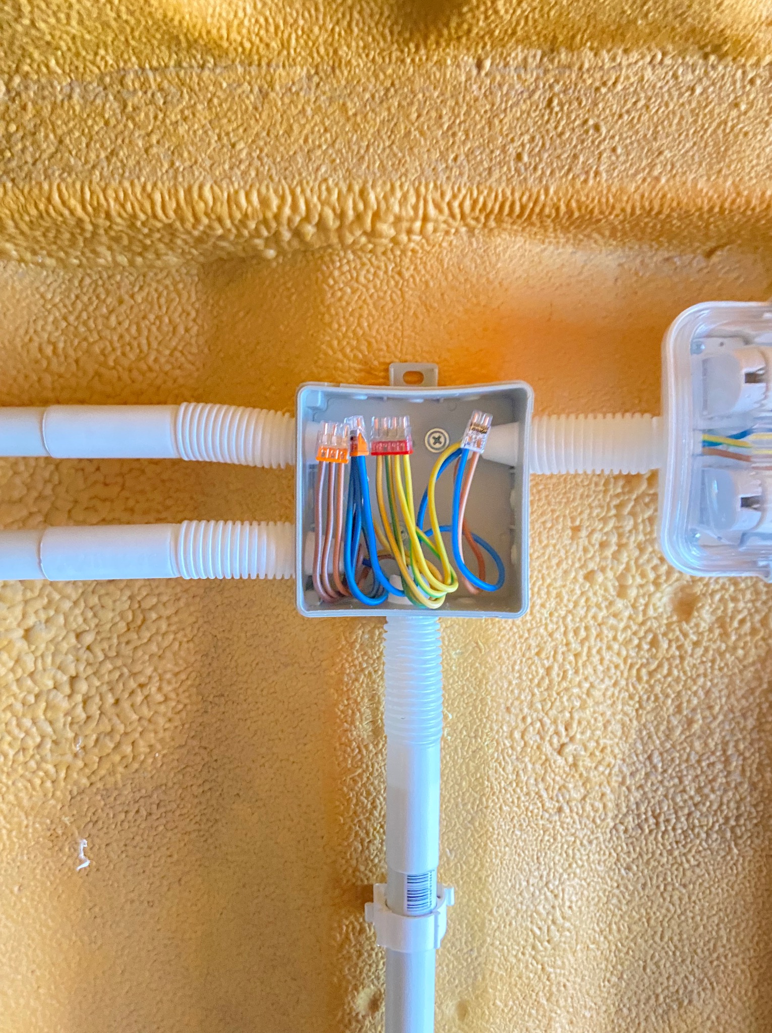 Połączenia przewodów w puszce instalacyjnej z widocznymi złączkami WAGO, umieszczonej na ścianie pokrytej pianką izolacyjną. Rury karbowane doprowadzają przewody.