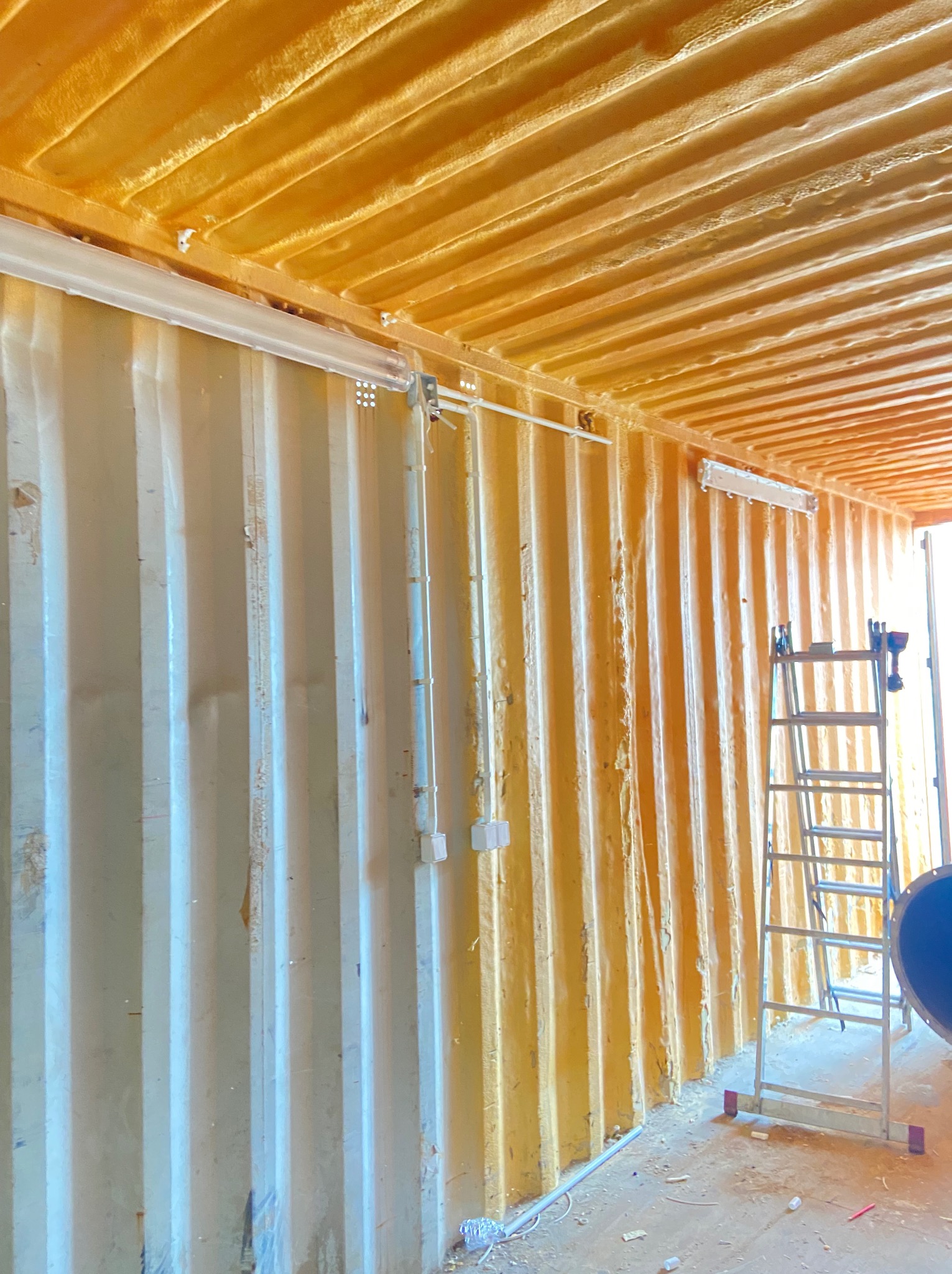 Wnętrze kontenera z ocieplonym stropem pianką PUR i zamontowanym oświetleniem. Widoczne przewody elektryczne i drabina. Zastosowano białe rury instalacyjne.