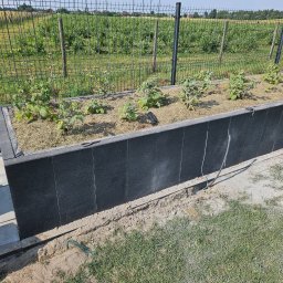 Mateusz Fraszczyński - Podwyższona rabata z czarnych paneli kompozytowych, wypełniona słomą i młodymi sadzonkami, otoczona metalową siatką ogrodzeniową na tle zieleni.