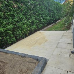 Mateusz Fraszczyński - Przygotowanie terenu pod ścieżkę z kostki brukowej: wyrównany piasek na części powierzchni, obrzeża z kostki betonowej, tło z wysokiego żywopłotu.