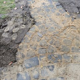 Mateusz Fraszczyński - Ścieżka z kostki granitowej w trakcie budowy, częściowo zasypana piaskiem, z widocznym fragmentem trawnika i błota po lewej stronie.