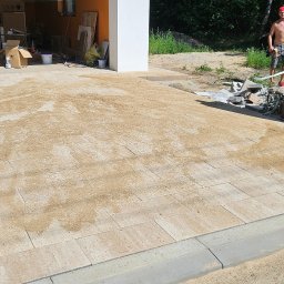 Mateusz Fraszczyński - Niedokończona nawierzchnia z kostki brukowej w Łodzi, częściowo zasypana piaskiem, widoczny obrzeże trawnika i fragment ogrodzenia z drewnianymi elementami.