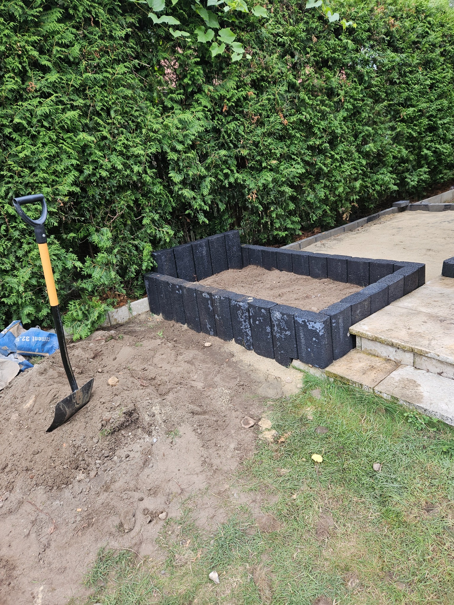 Przygotowanie rabaty kwiatowej z wykorzystaniem czarnej kostki brukowej, widoczna łopata i worki z cementem, w tle wysokie tuje.