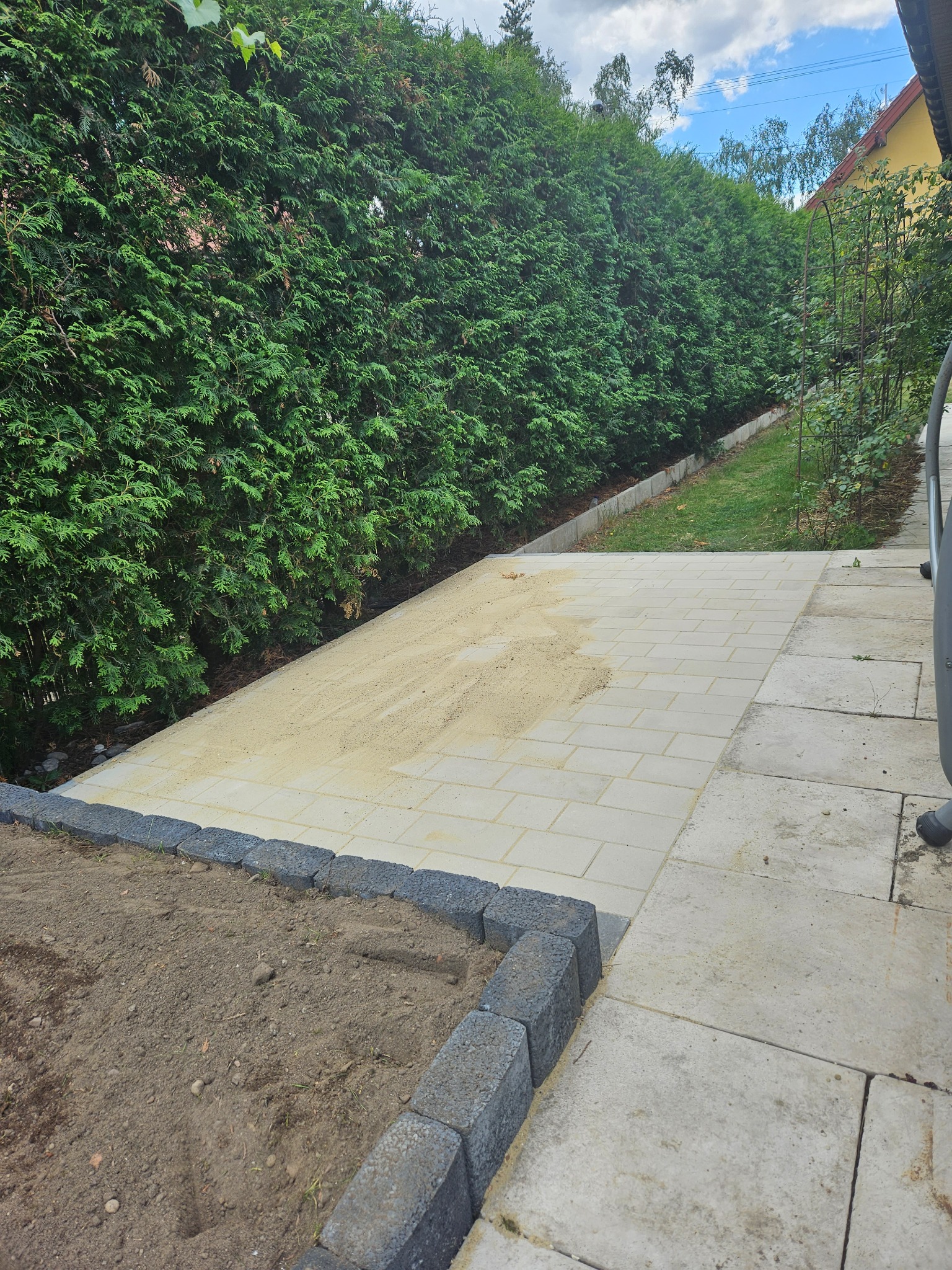 Przygotowanie terenu pod ścieżkę z kostki brukowej: wyrównany piasek na części powierzchni, obrzeża z kostki betonowej, tło z wysokiego żywopłotu.