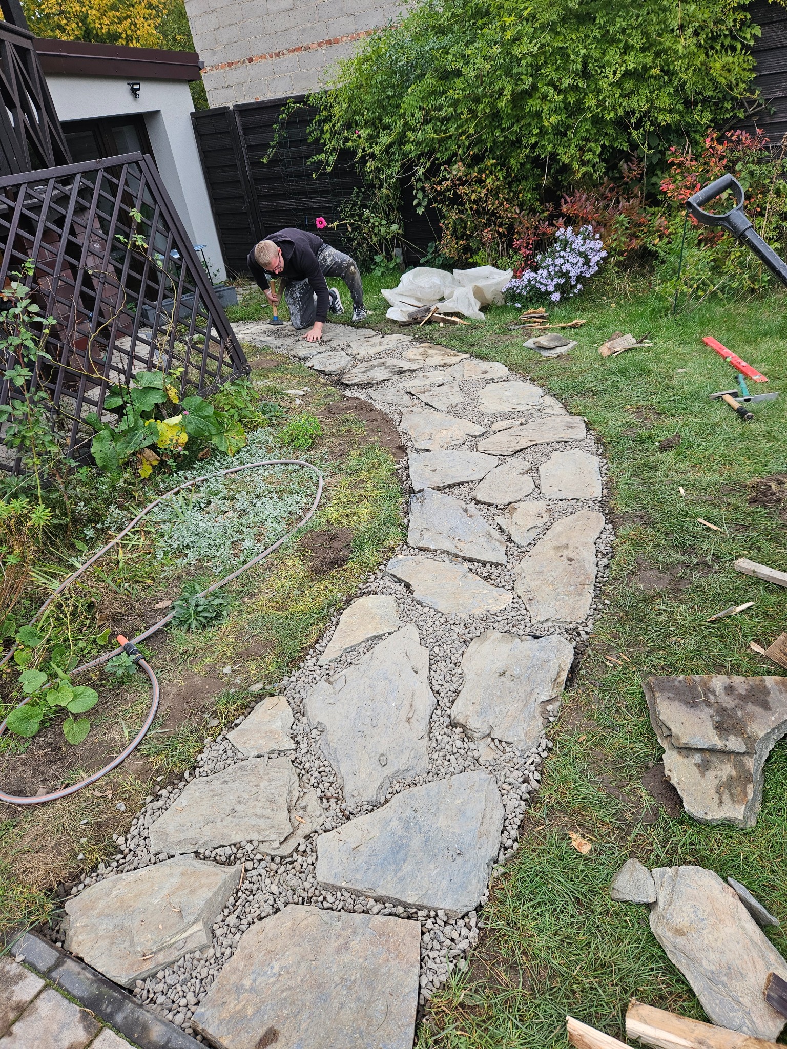 Pracownik w trakcie tworzenia ścieżki z nieregularnych płyt kamiennych, wypełnionych drobnym żwirem, w ogrodzie z widocznymi narzędziami i roślinnością.
