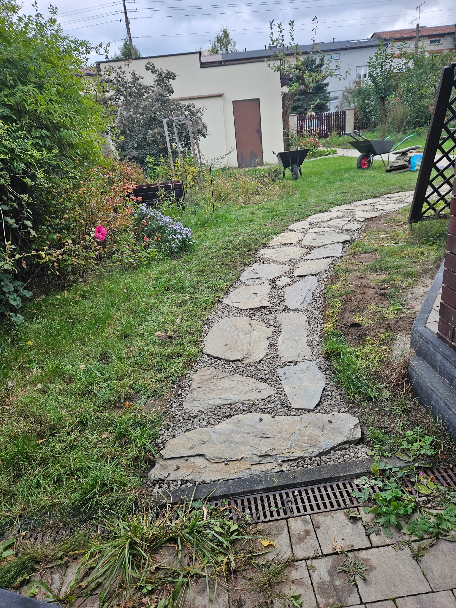 Ścieżka z nieregularnych płyt kamiennych, wysypana drobnym żwirem, prowadząca przez trawnik w ogrodzie, zakończona kratką drenażową