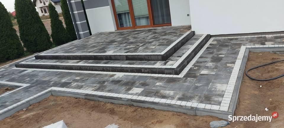 Schody wejściowe do domu wyłożone kostką brukową w odcieniach szarości, z białymi obrzeżami, widoczne niedokończone prace ziemne obok.