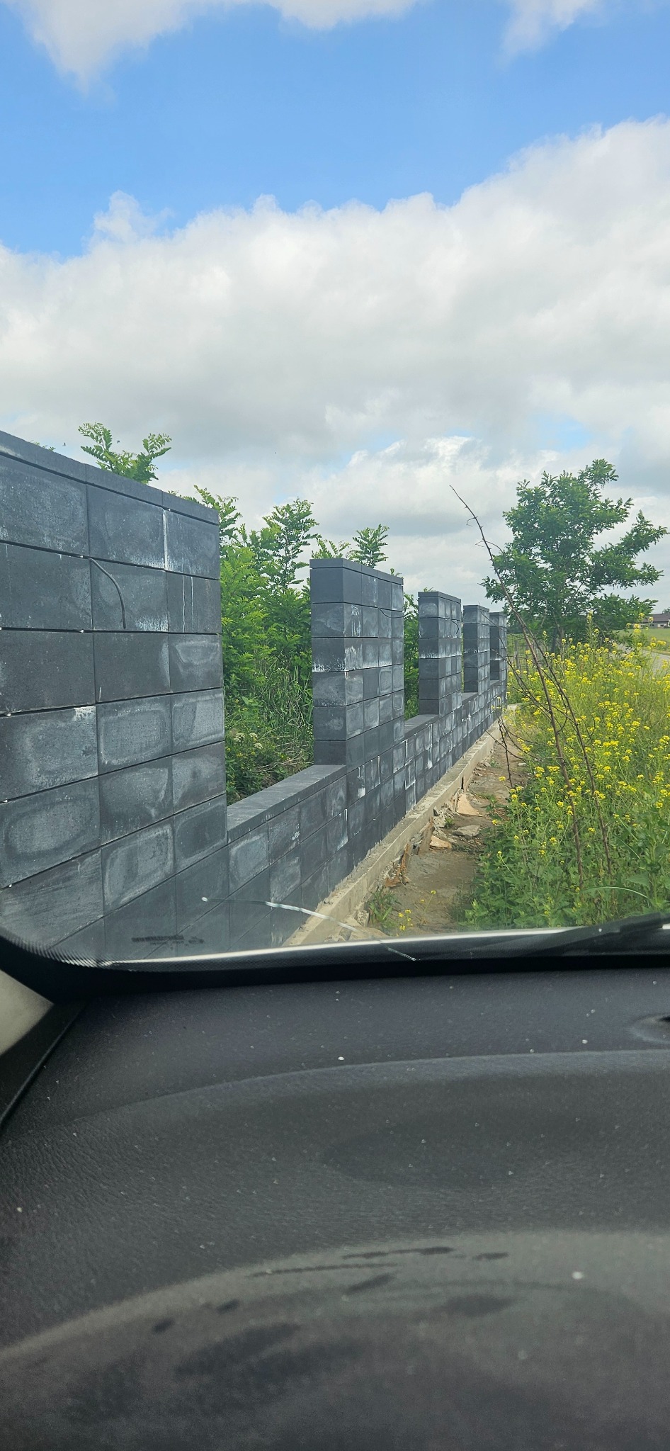 Szary, modułowy mur ogrodzeniowy z pustaków betonowych, widok zza szyby samochodu, z żółtymi kwiatami i zielenią w tle.