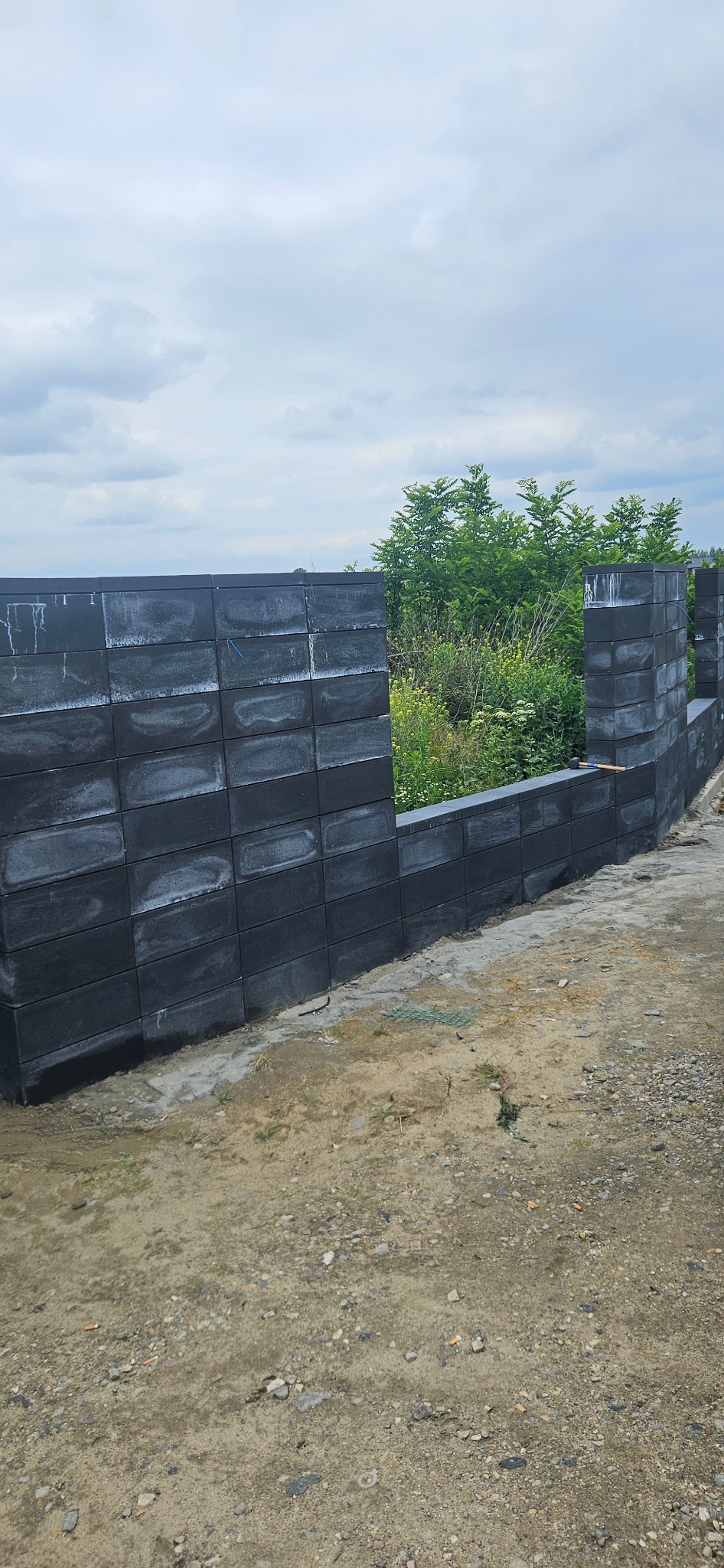 Wznoszenie muru z szarych bloczków betonowych, widoczny fragment ogrodzenia w trakcie budowy, młotek na górnej krawędzi muru, w tle zieleń.