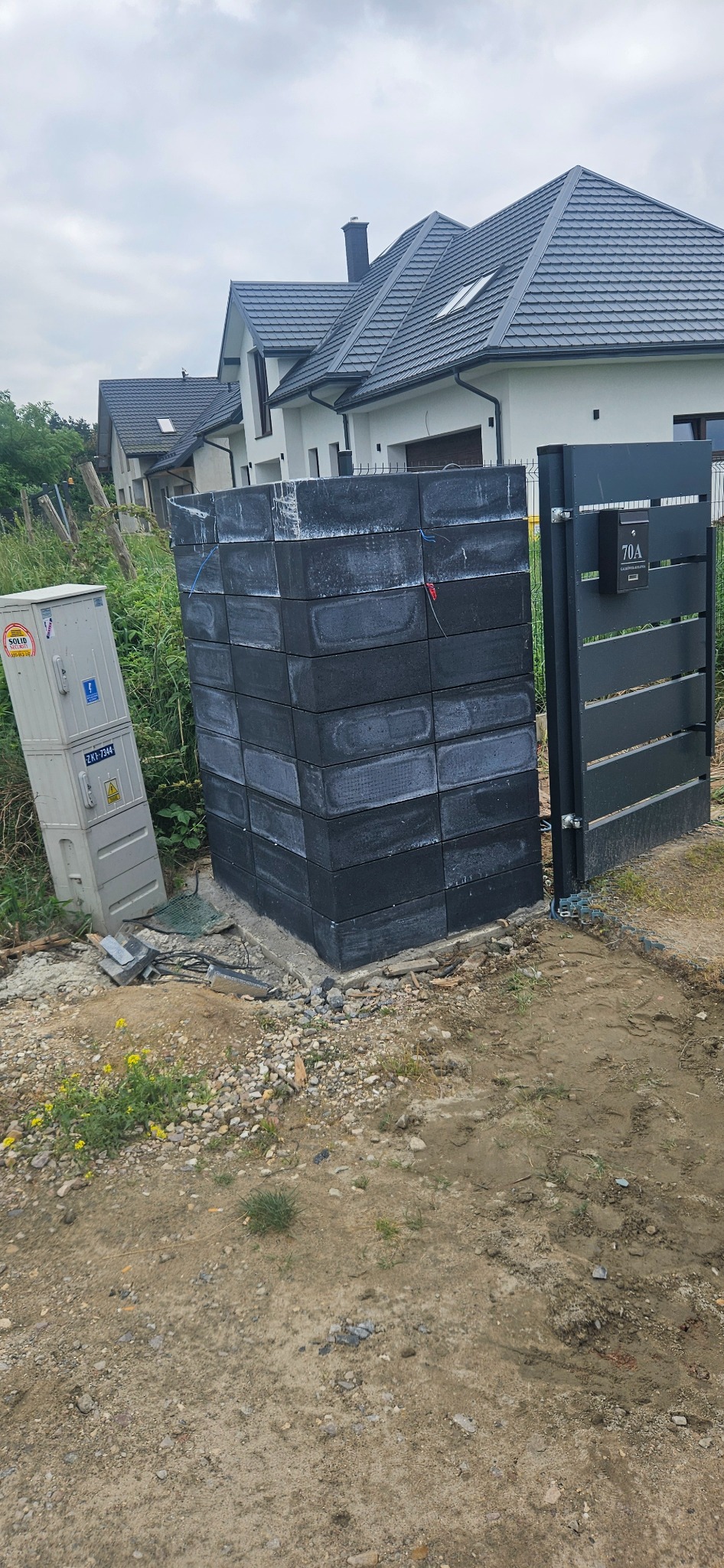Stos szarych bloczków betonowych ułożonych obok metalowej furtki z numerem domu 70A, widoczna skrzynka elektryczna i fragment posesji z domem w tle.