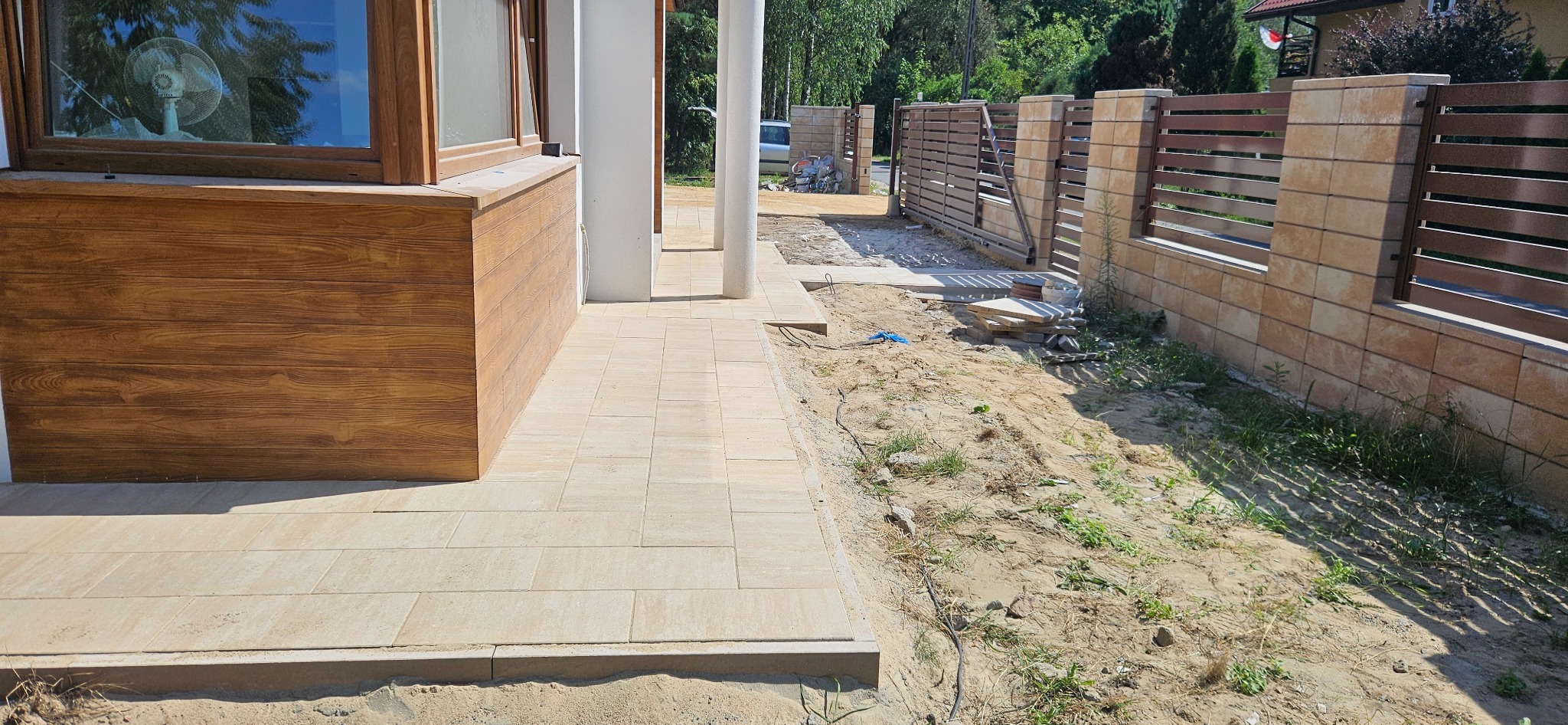 Jasne, prostokątne płyty chodnikowe ułożone przed budynkiem z drewnianą elewacją i białymi kolumnami, w tle widoczny fragment ogrodzenia z poziomych, brązowych desek i kamiennych słupków.