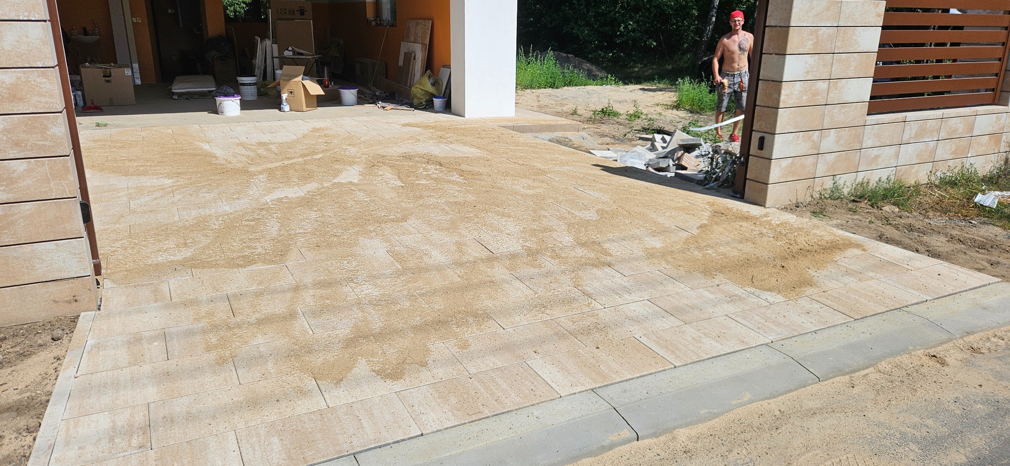 Niedokończona nawierzchnia z kostki brukowej w Łodzi, częściowo zasypana piaskiem, widoczny obrzeże trawnika i fragment ogrodzenia z drewnianymi elementami.