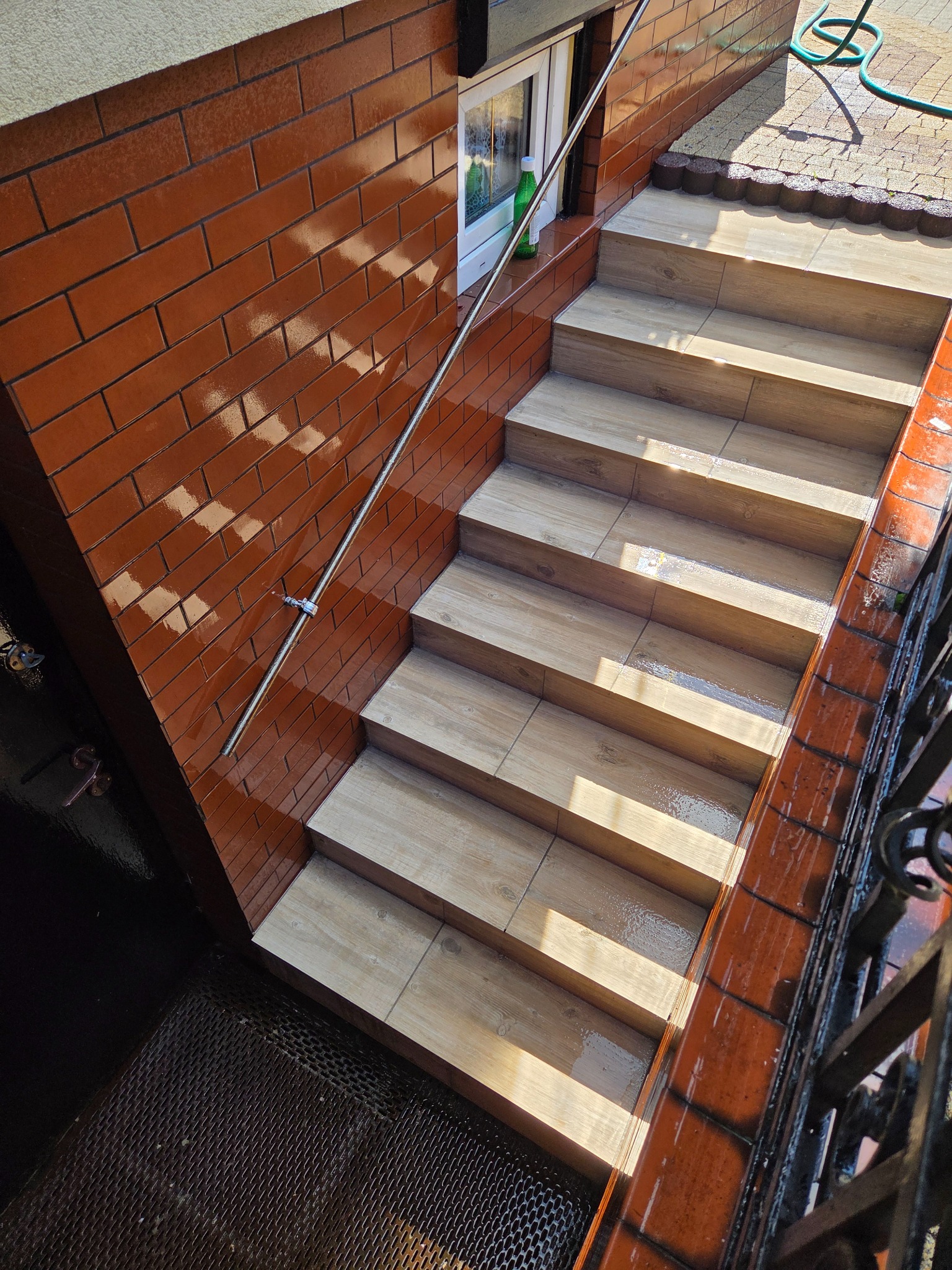 Wykończone schody zewnętrzne z drewnopodobnych płytek, prowadzące do drzwi. Widoczna ceglana ściana i metalowa barierka. Całość w ciepłej kolorystyce.