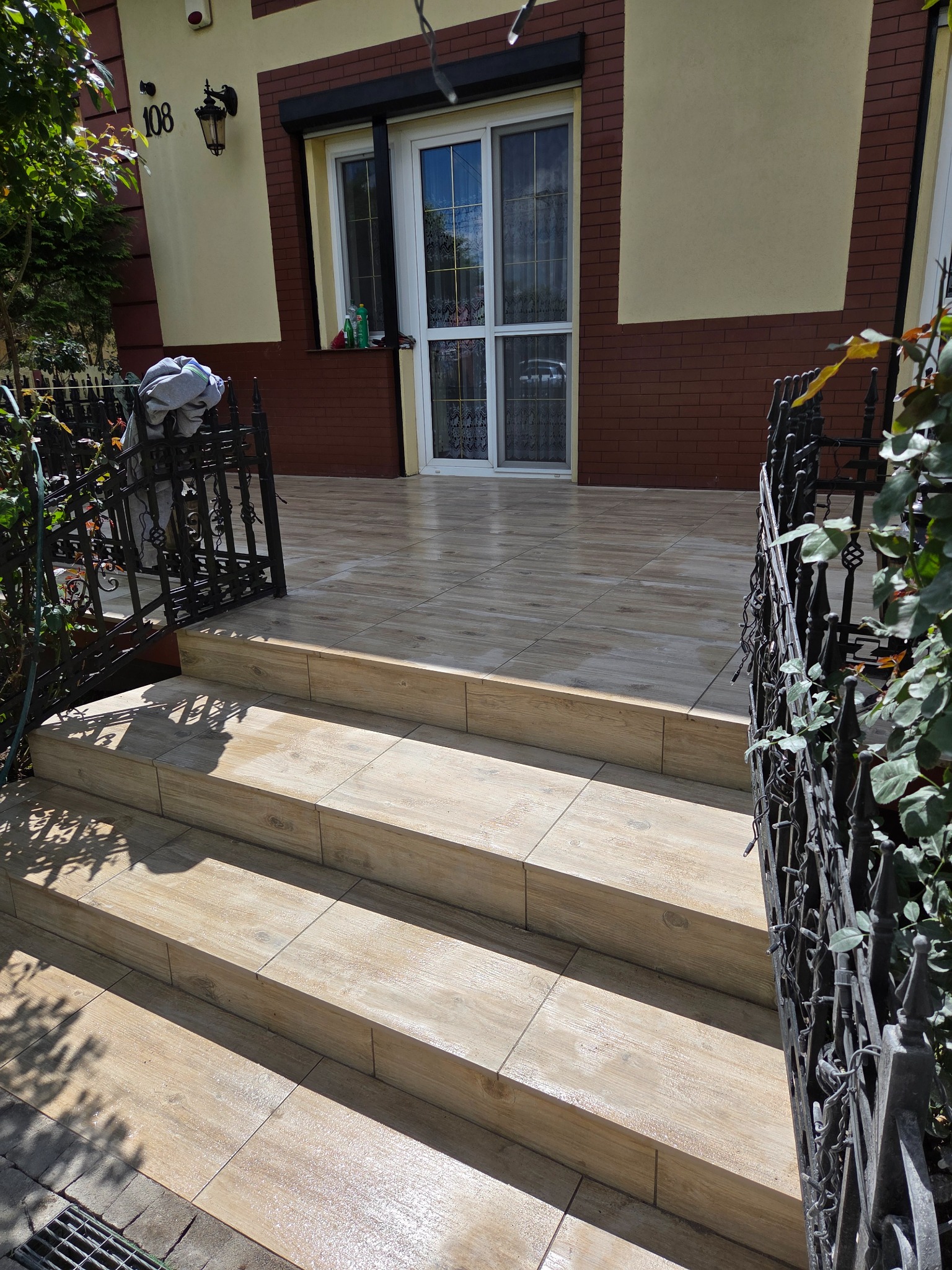 Kafelkowane schody wejściowe do domu z kutą balustradą. Jasne płytki imitujące drewno prowadzą na taras z brązowymi płytkami. Widok na elewację domu.