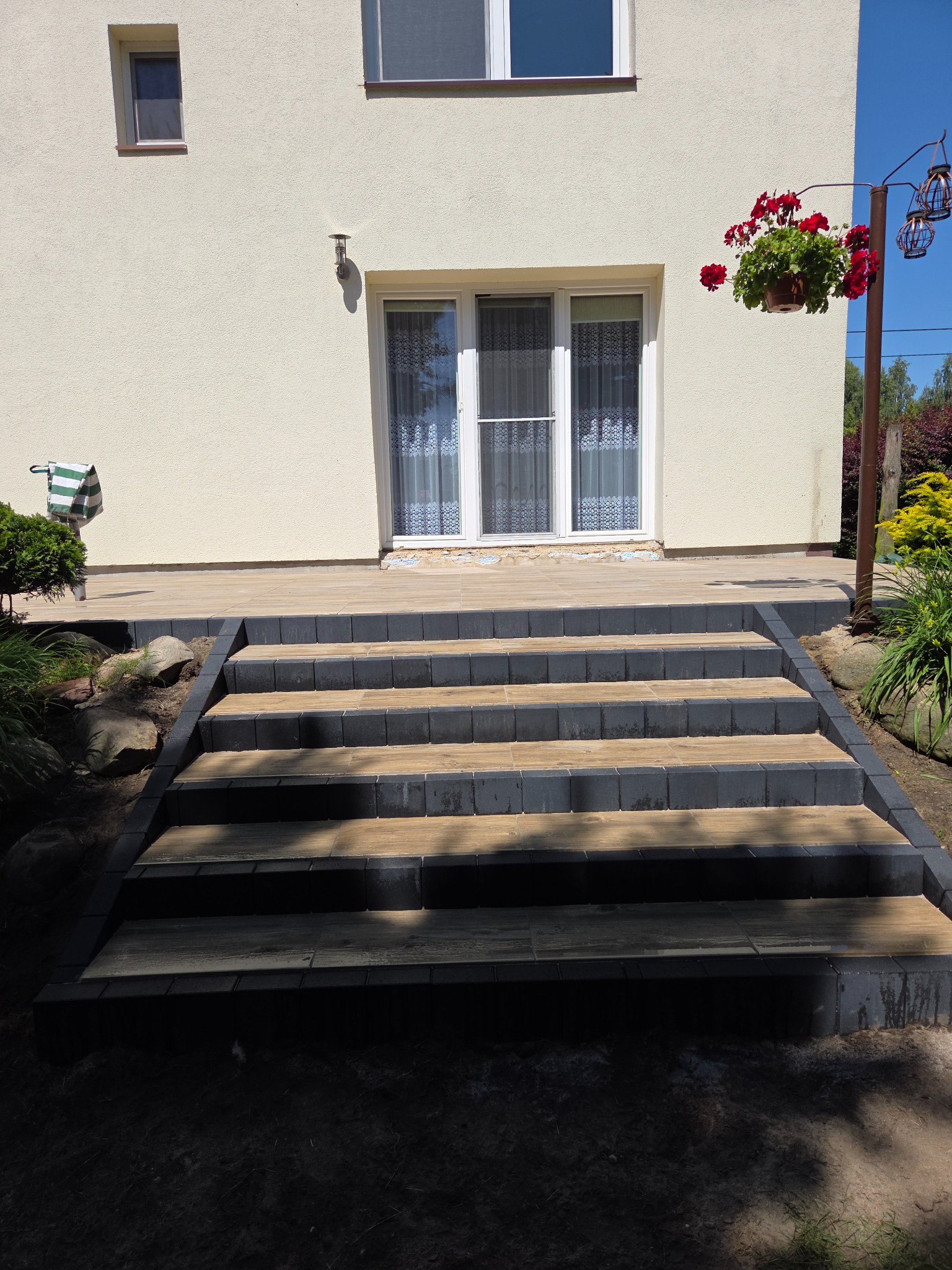 Schody wejściowe z kostki brukowej w odcieniach brązu i grafitu prowadzące do domu z kremową elewacją, ozdobione zielenią i elementami dekoracyjnymi.