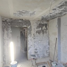 MasterOK OLEH VASYLOVSKYI - Wnętrze w trakcie remontu: ściany z resztkami starej farby, narzędzia i materiały budowlane, widoczna postać w przejściu. Przygotowanie do szpachlowania.
