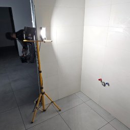 MasterOK OLEH VASYLOVSKYI - Wnętrze łazienki w trakcie wykończenia: białe płytki ścienne, szare płytki podłogowe, widoczne przyłącza wodne. W tle pracownik i lampa budowlana.