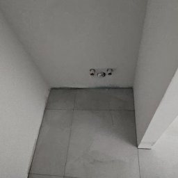 MasterOK OLEH VASYLOVSKYI - Szare, kwadratowe płytki podłogowe w małym pomieszczeniu, widoczne fragmenty stóp w obuwiu. Na ścianie widoczne trzy otwory instalacyjne.