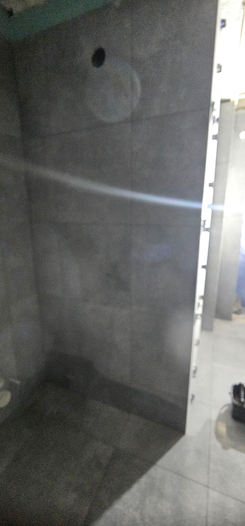 Wykończona, ciemnoszara kabina prysznicowa z widocznym odpływem i otworem na słuchawkę. Minimalistyczny design, płytki o jednolitej fakturze, jasne fugi.