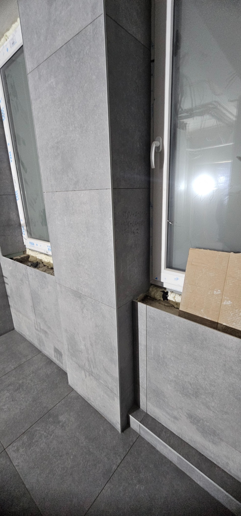 Szare płytki imitujące kamień na ścianie i podłodze w łazience, widoczne okno z białymi ramami i fragment instalacji. Minimalistyczny design, nowoczesne wykończenie.