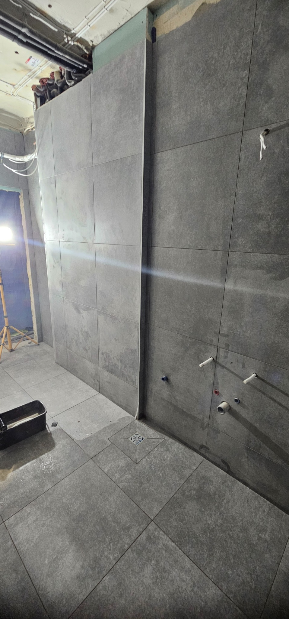 Wykończona łazienka z szarymi, kwadratowymi płytkami na ścianach i podłodze. Widoczny odpływ liniowy i wystające rury. W tle lampa budowlana.