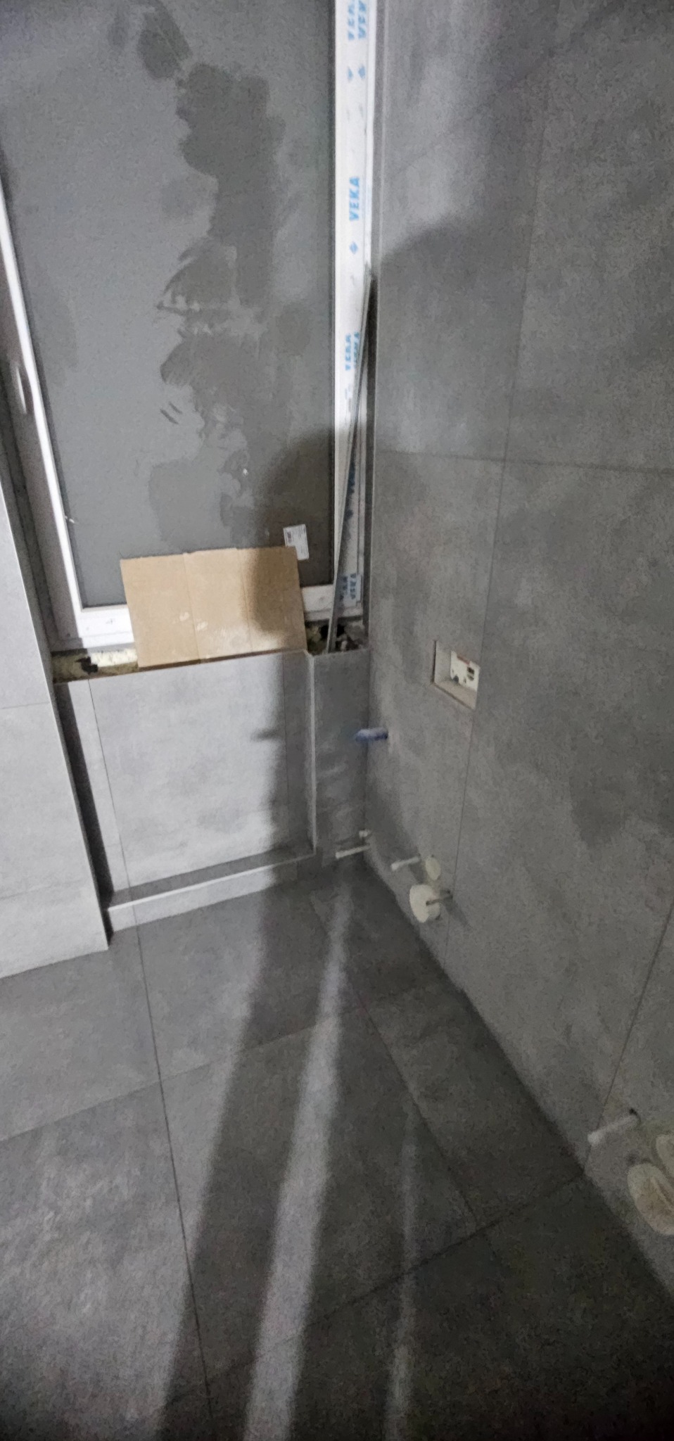 Szare płytki podłogowe i ścienne w trakcie wykańczania łazienki. Widoczne fragmenty instalacji wodnej i okna. Minimalistyczny design w surowym wydaniu.