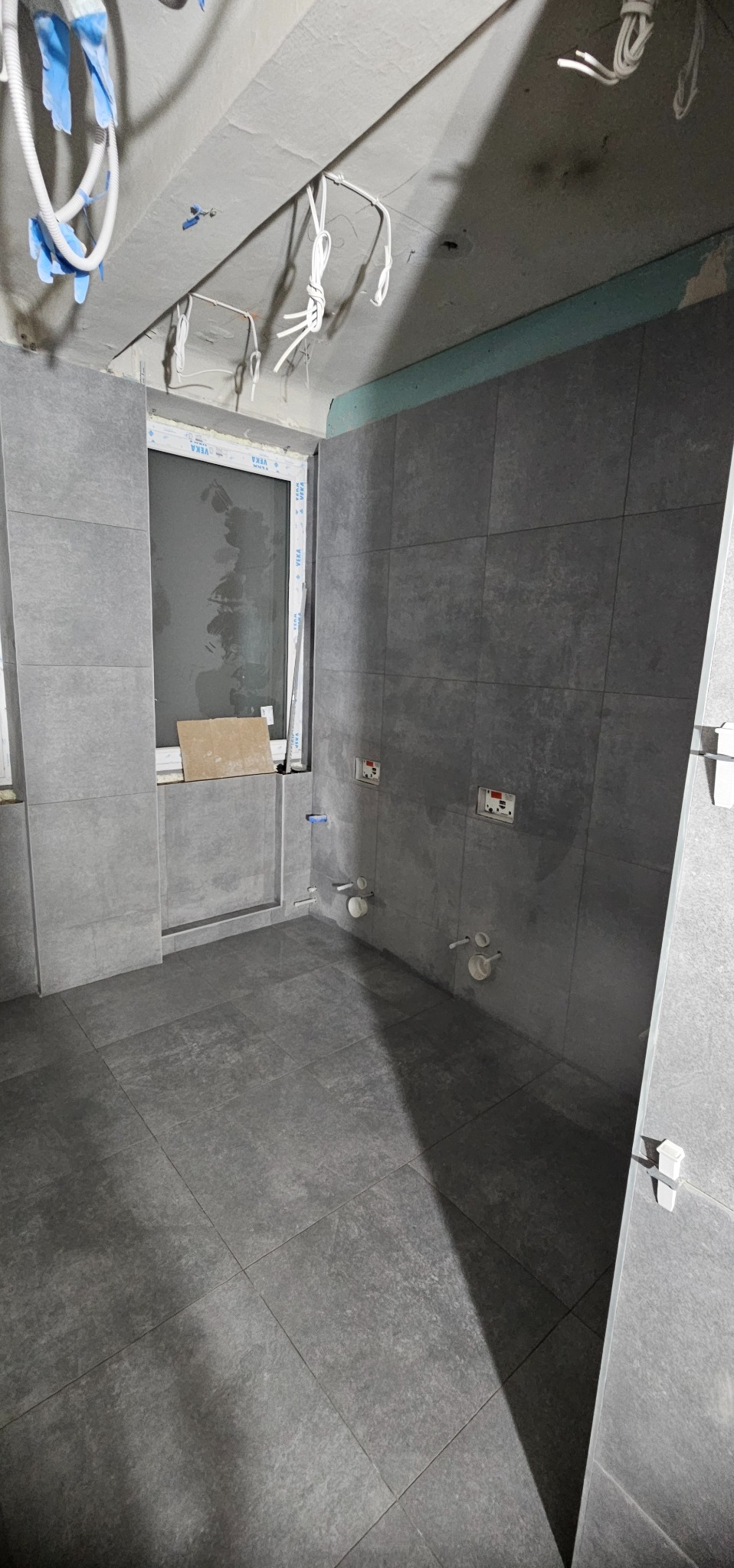 Szare płytki podłogowe i ścienne w surowej łazience z widocznymi instalacjami. Okno z parapetem. Białe przewody zwisają z sufitu. Etap wykończenia.