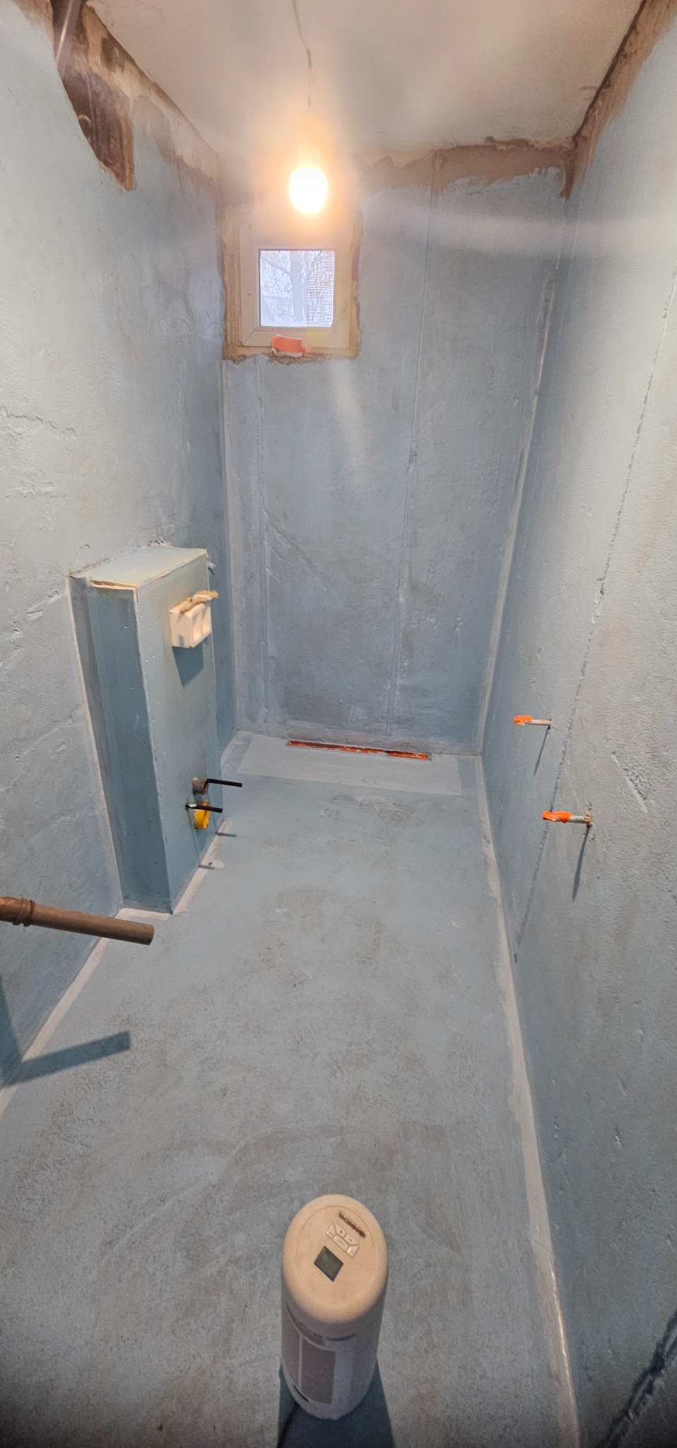 Niebieskie wnętrze łazienki po hydroizolacji, widoczne rury, okno i lampa. Przygotowanie do wykończenia, z bliska widoczny czujnik wilgotności.