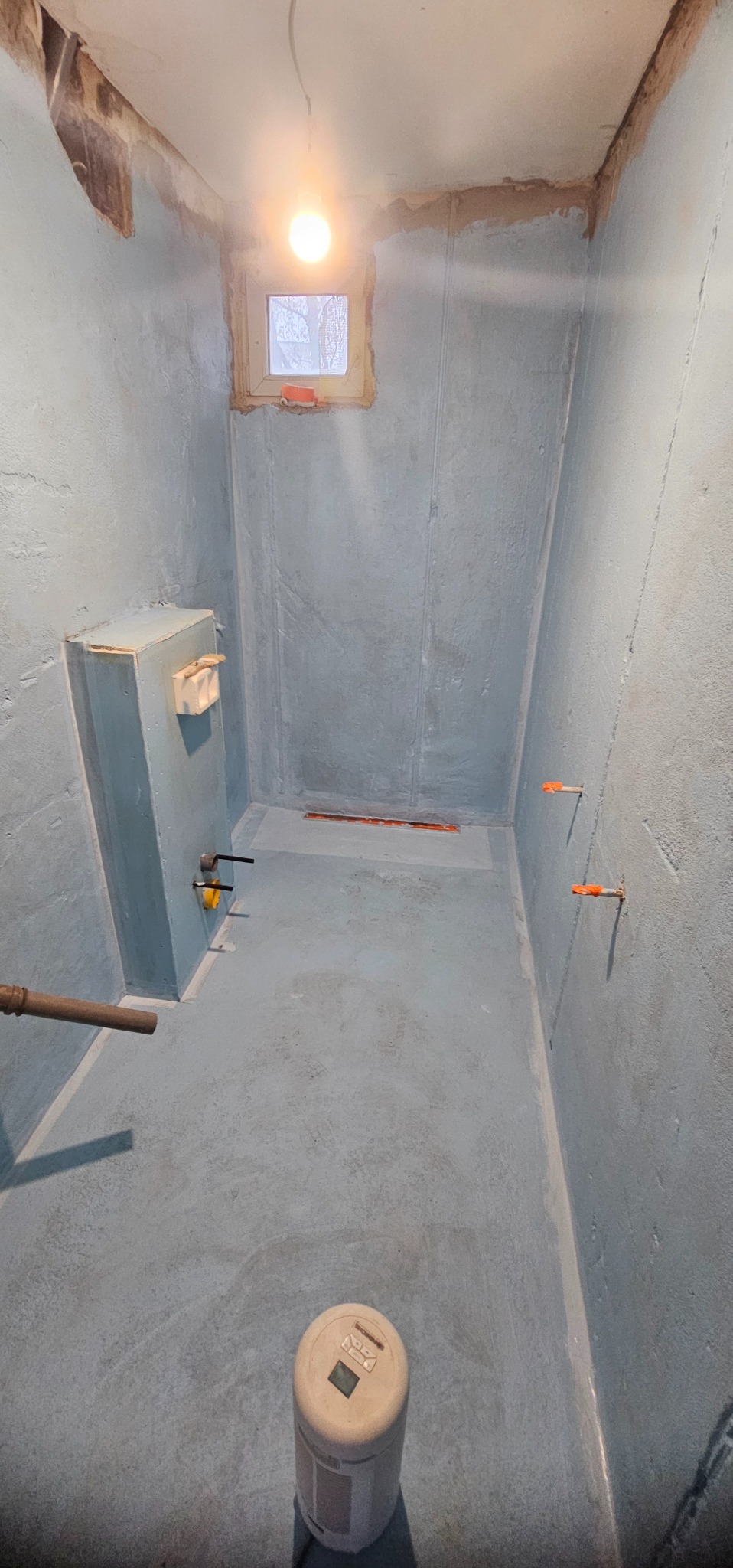 Błękitne wnętrze łazienki w trakcie remontu. Widoczne przygotowane ściany, instalacje i odpływ liniowy. Małe okno z widokiem na zewnątrz.