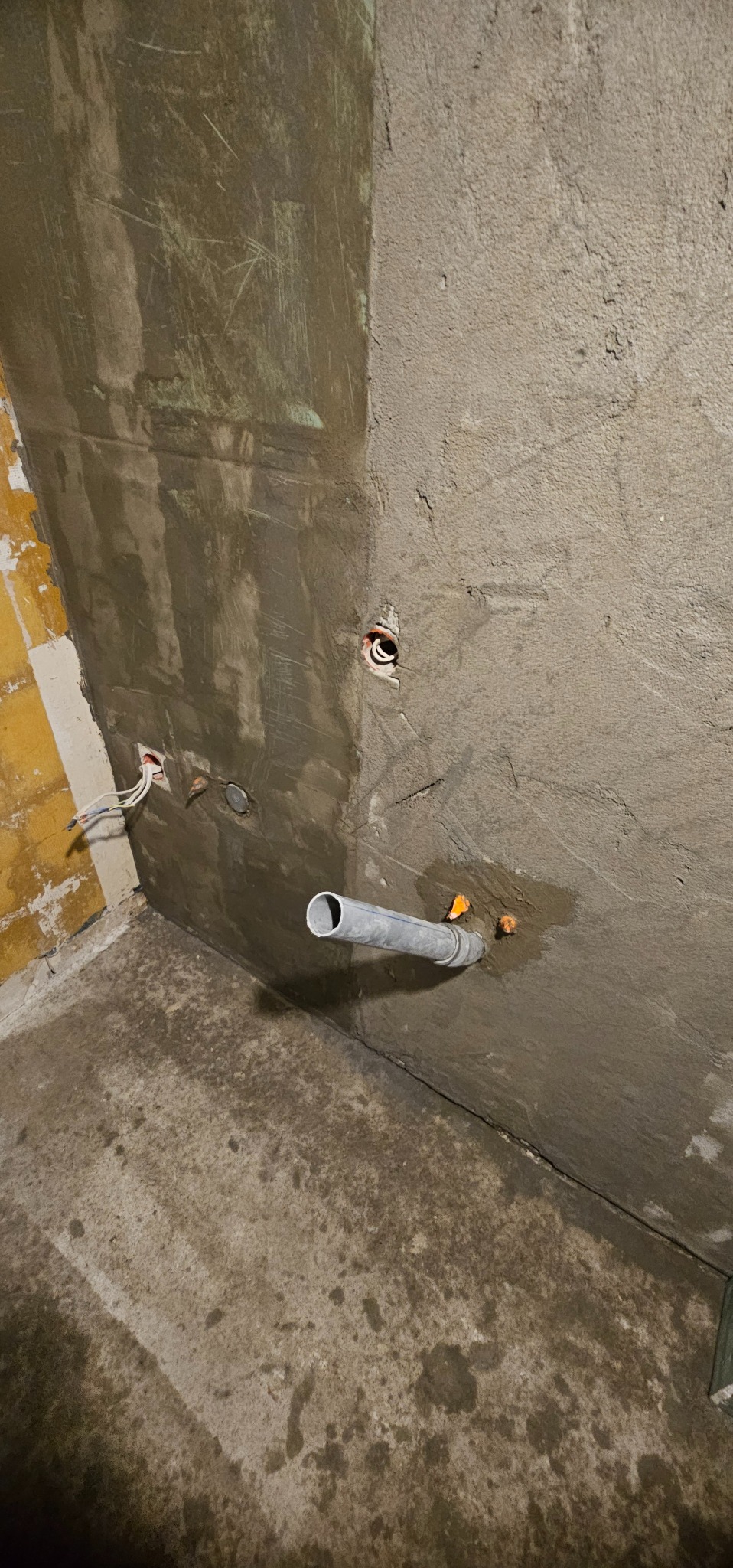 Ściana w trakcie szpachlowania z wystającą rurą i przewodami elektrycznymi. Widoczne ślady wilgoci na podłodze i fragment żółtej ściany w rogu.
