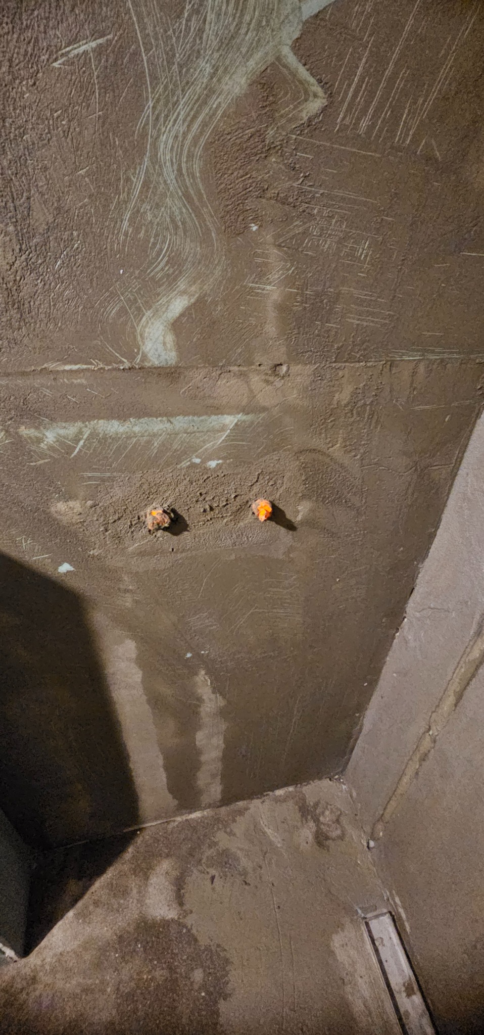 Świeżo nałożona, brązowa warstwa szpachli na ścianie w rogu pomieszczenia, widoczne ślady po szpachlowaniu i dwa wystające elementy instalacji.