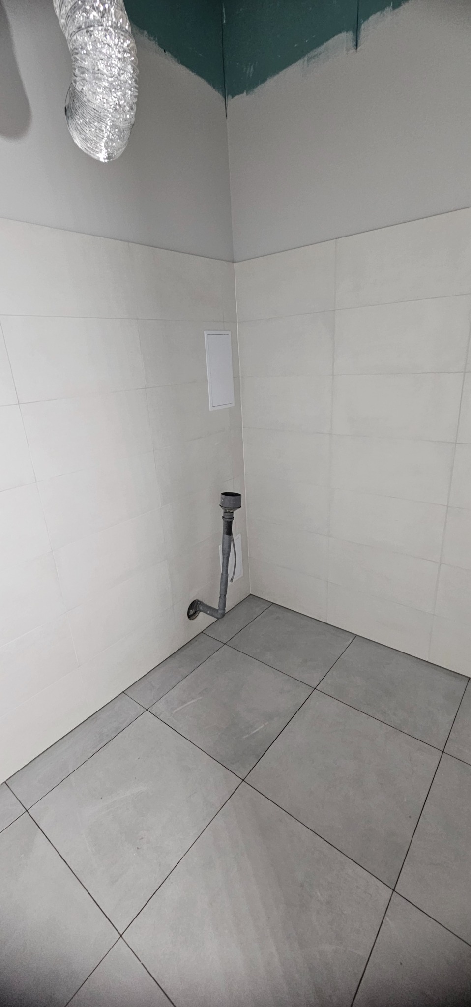 Wykończona łazienka z szarymi płytkami podłogowymi i białymi ściennymi. Widoczny fragment instalacji kanalizacyjnej i rura wentylacyjna. Górna część ściany pomalowana na zielono.