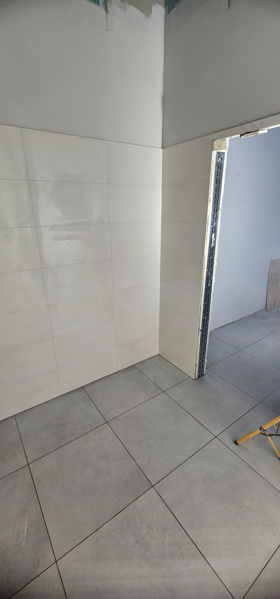 Wykończona ściana z błyszczących, białych płytek w łazience. Na podłodze szare płytki. Widoczny fragment ościeżnicy drzwiowej i lampa robocza.