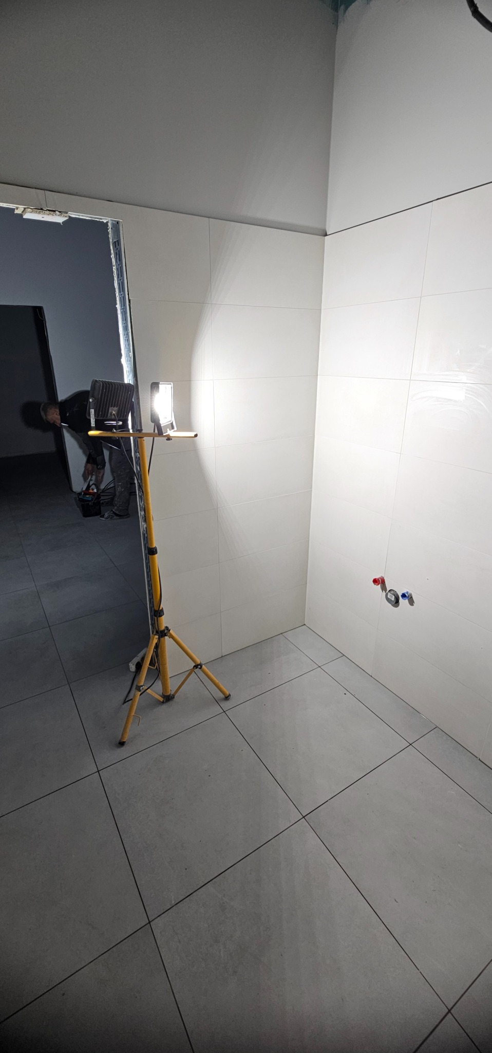 Wnętrze łazienki w trakcie wykończenia: białe płytki ścienne, szare płytki podłogowe, widoczne przyłącza wodne. W tle pracownik i lampa budowlana.