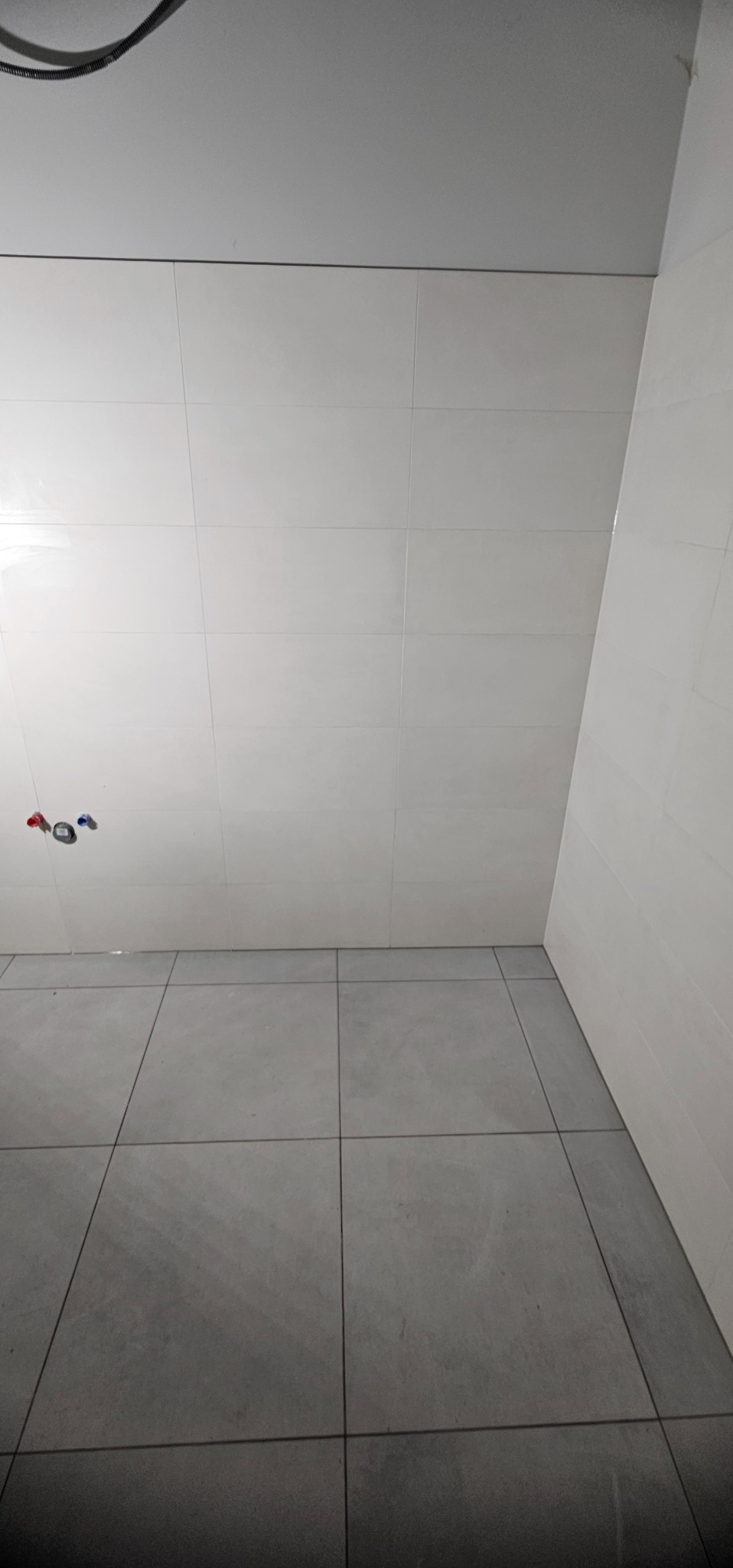 Wykończona łazienka z białymi płytkami na ścianach i szarymi na podłodze. Widoczne przyłącza wodne. Minimalistyczny design, czyste linie, nowoczesne wykończenie.