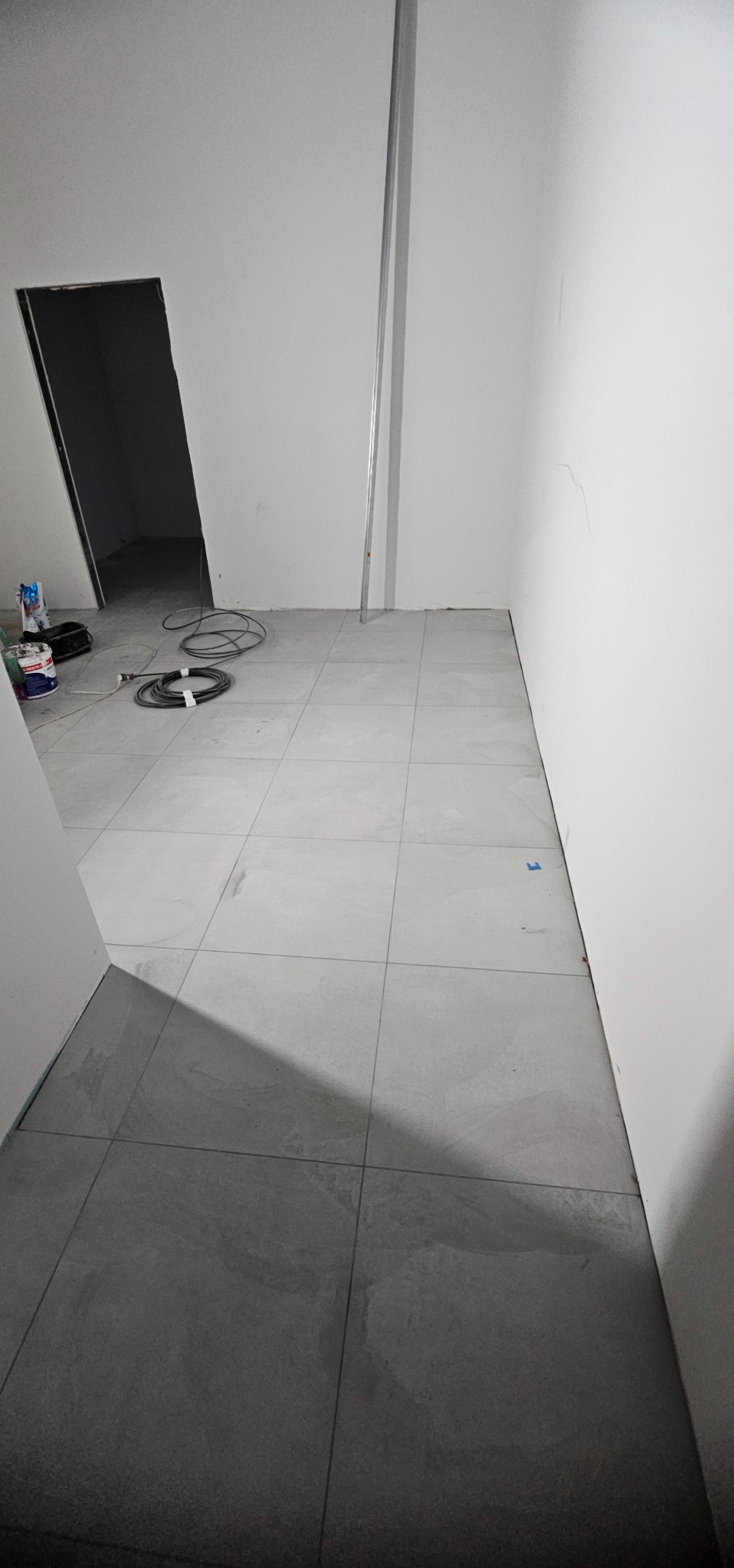 Jasne, kwadratowe płytki podłogowe w surowym wnętrzu. Widoczne puszki z farbą, kable i metalowy profil przy ścianie. Minimalistyczny design, neutralna kolorystyka.